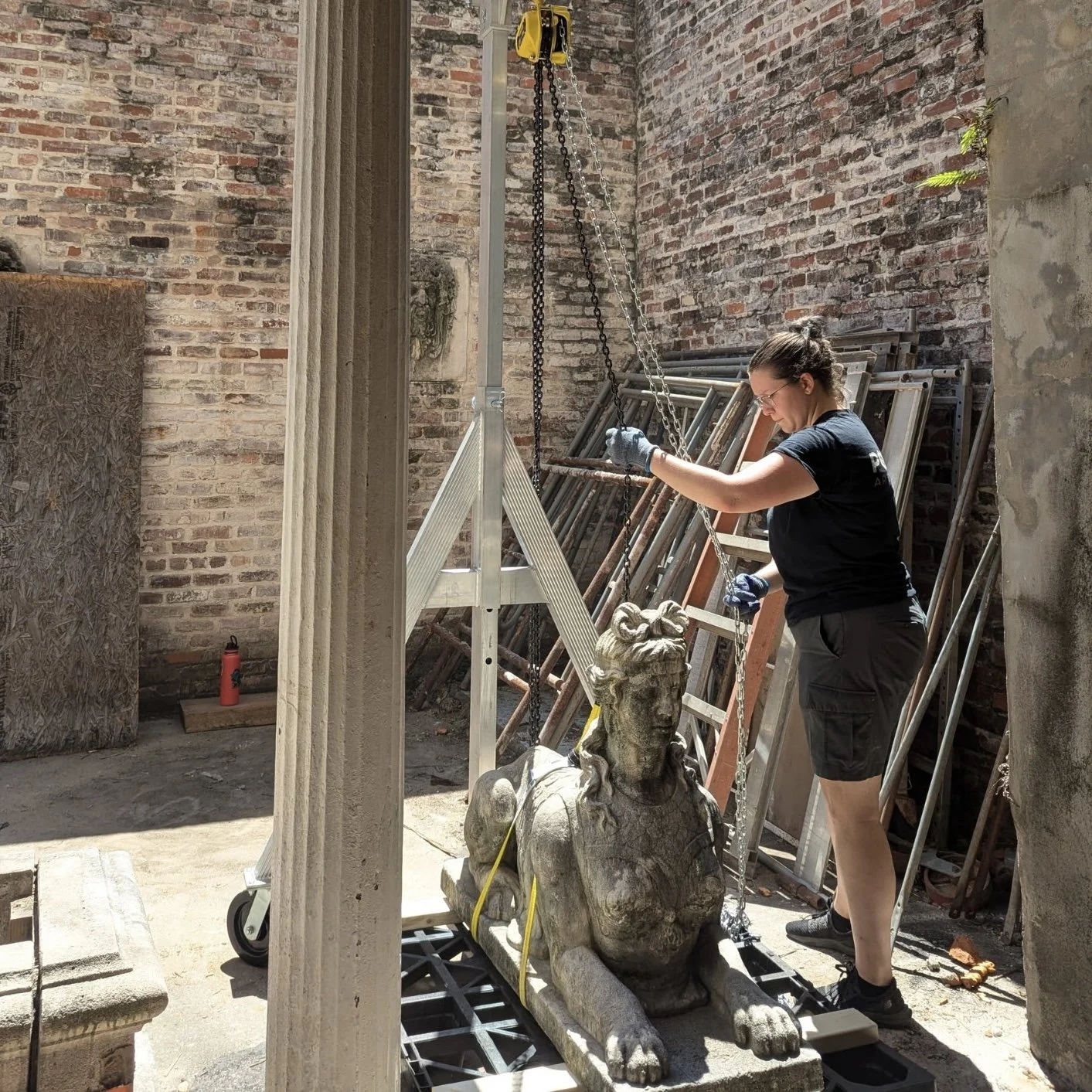 A woman working on a sculpture of a lion in a warehouse or workshop with brick walls and various frames and objects in the background.