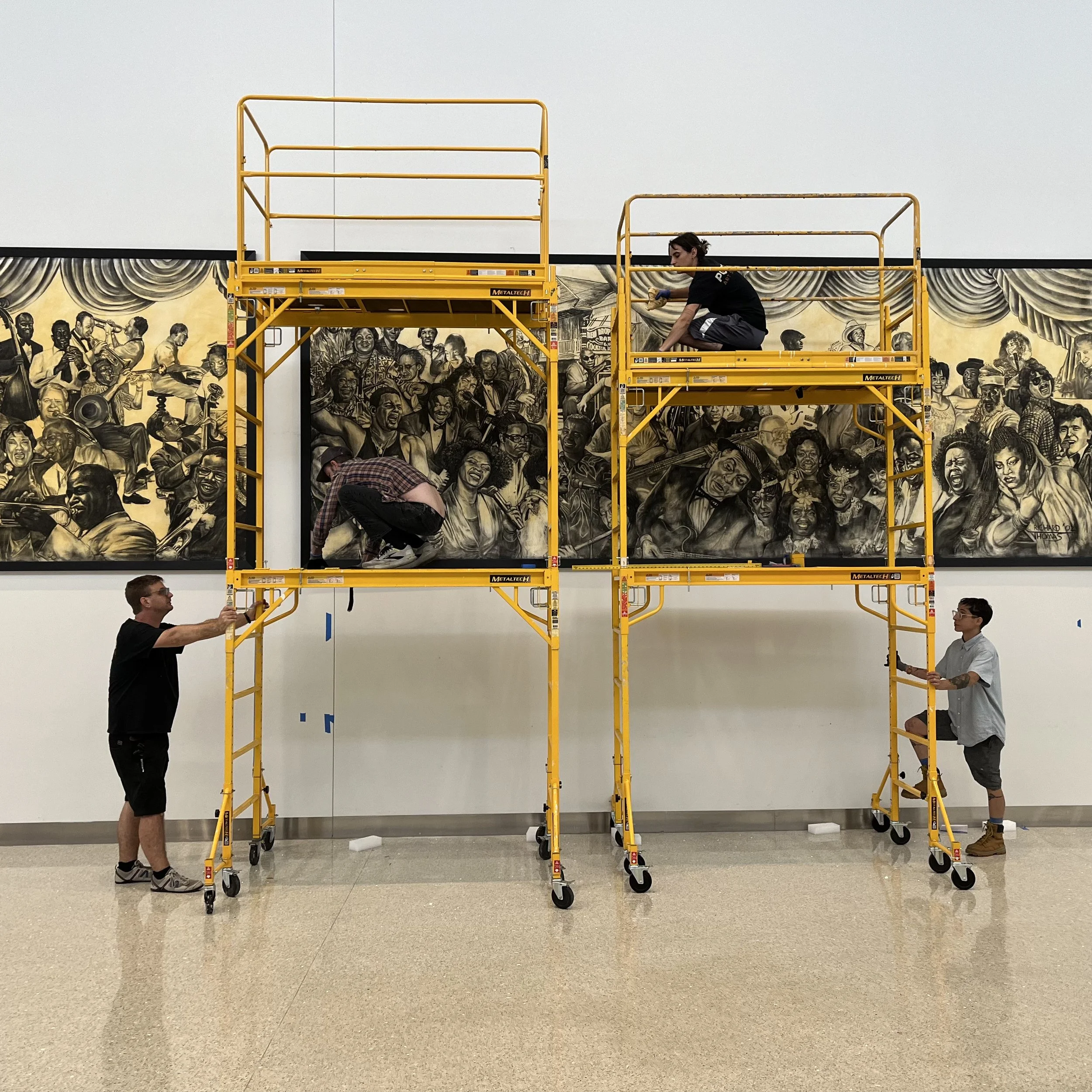 Two people assemble a large yellow scaffolding in front of a mural of a black and white jazz band on a wall inside a building.