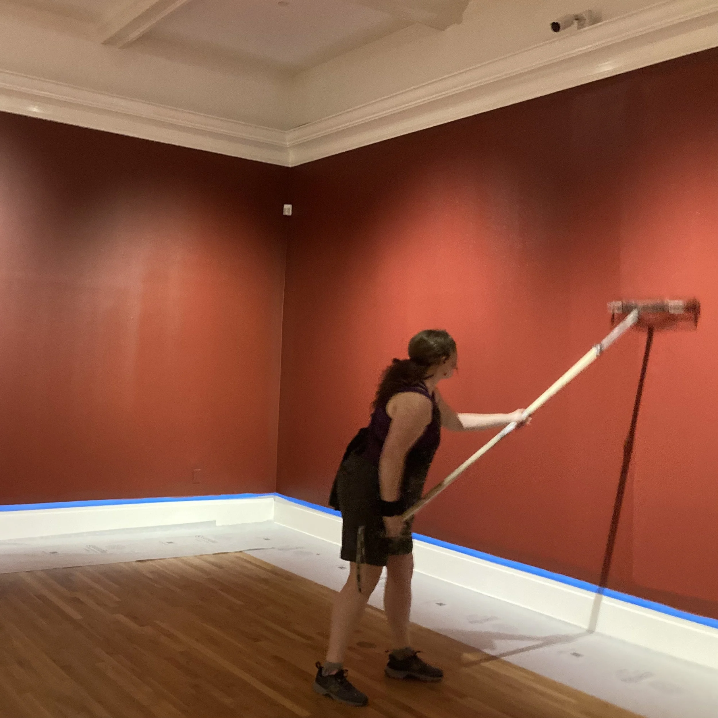 A person standing in a room with red walls, hardwood floor, and crown molding on the ceiling. The person's legs and shoes are visible, but their upper body and face are obscured by unseen objects.