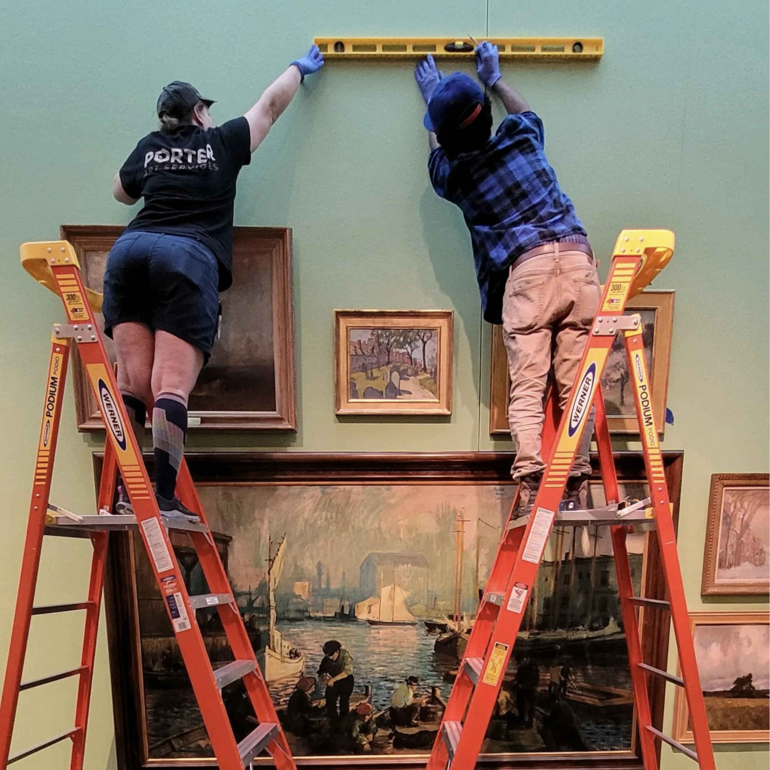 Two people on ladders hanging a framed painting of a scenic waterfront with boats and buildings on a green wall.