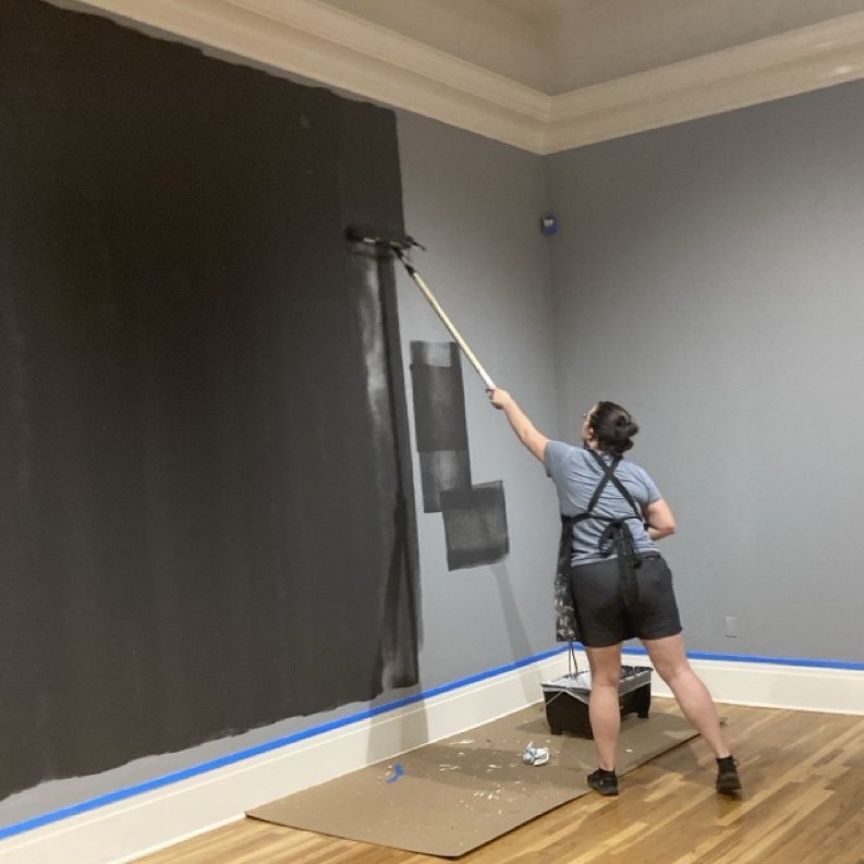 A person painting a museum wall black in a room, standing on the floor, using a long-handled paint roller.