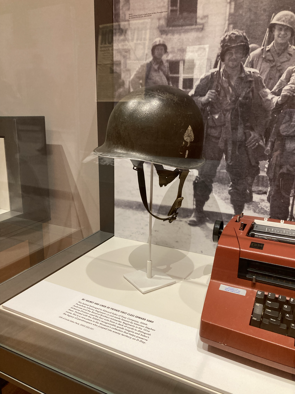 Military helmet and a typewriter on display in a museum exhibit with a black-and-white photo of soldiers in the background.