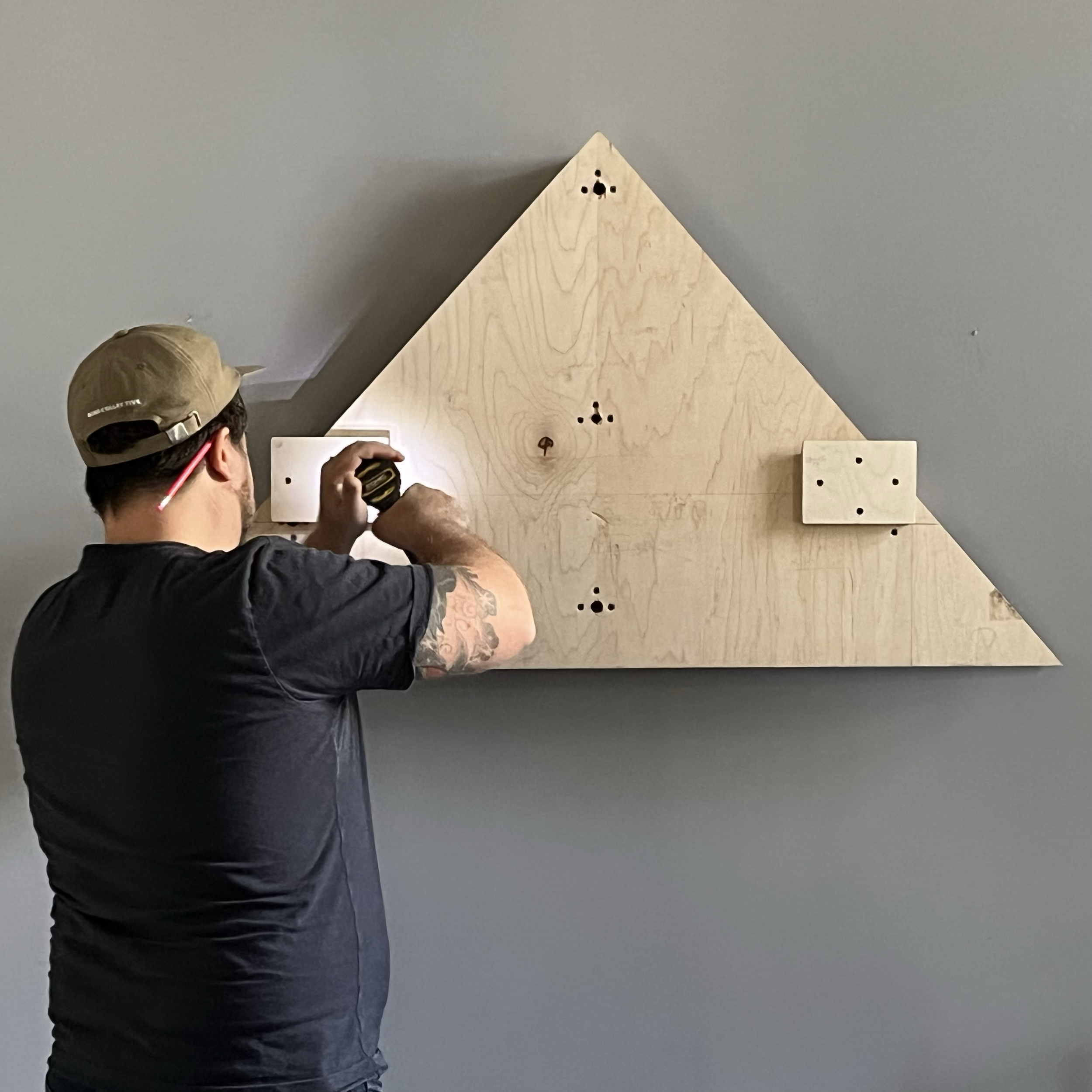Person with a baseball cap and glasses assembling a wooden mount that will later hold an art piece against a gray wall.