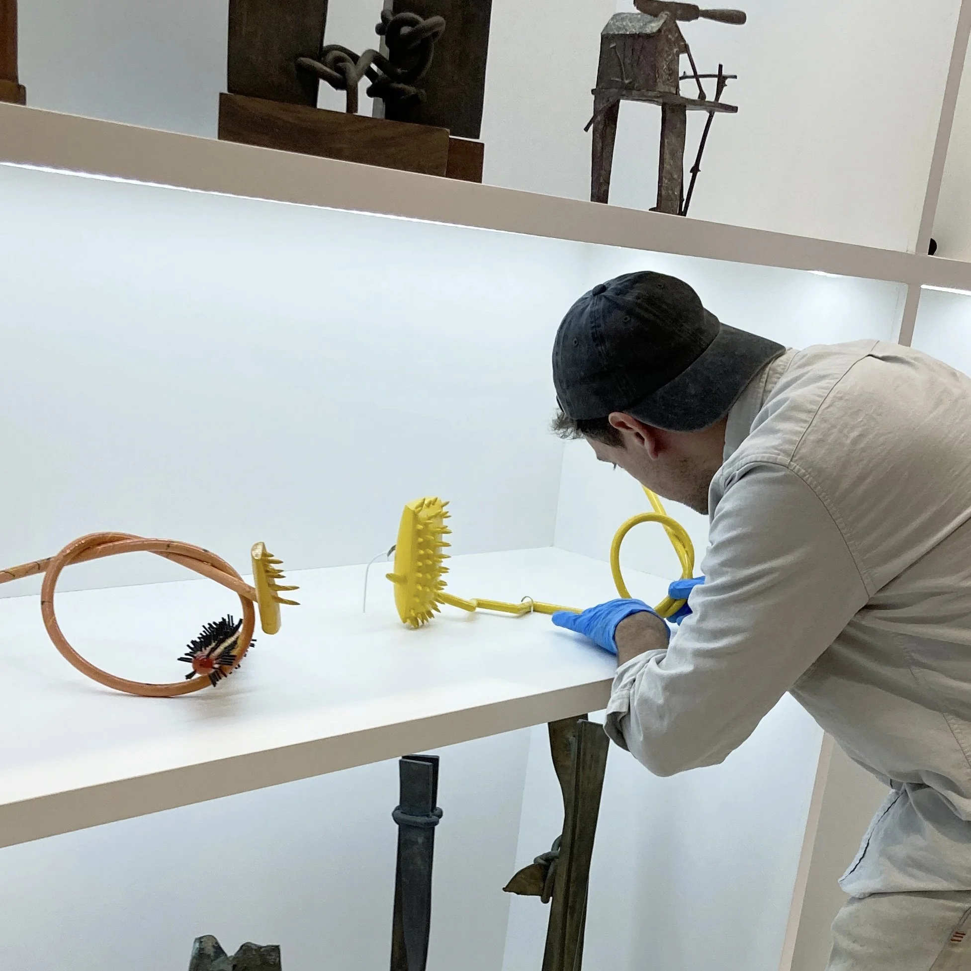 Person wearing a dark baseball cap, a white jacket, and blue gloves working on a craft project at a white table with various textured objects, including a yellow gear-shaped item and a black knurled knob.