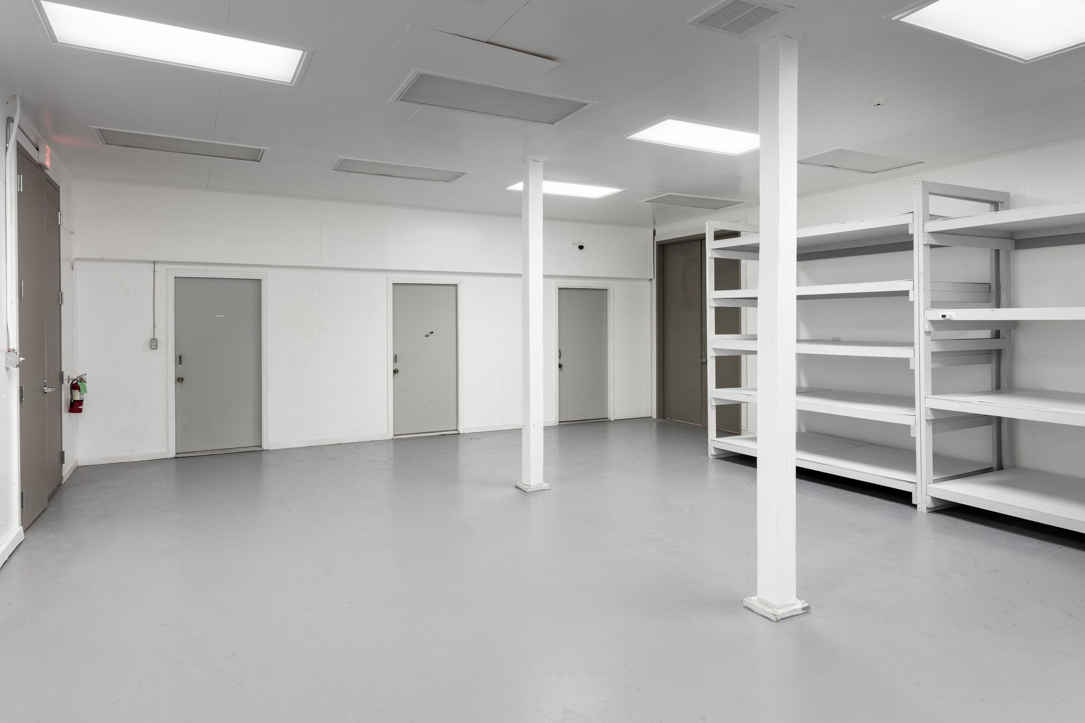 Empty commercial or storage room with white walls and ceiling, fluorescent lights, gray doors, and white metal shelving units.