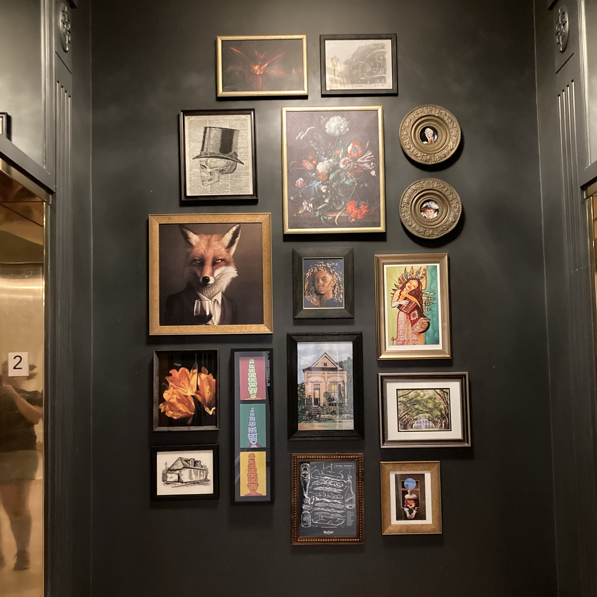 A dark wall decorated with various framed artwork, including a picture of a fox wearing a suit and tie, a colorful floral painting, a skeleton with a top hat, and several other art pieces and portraits.