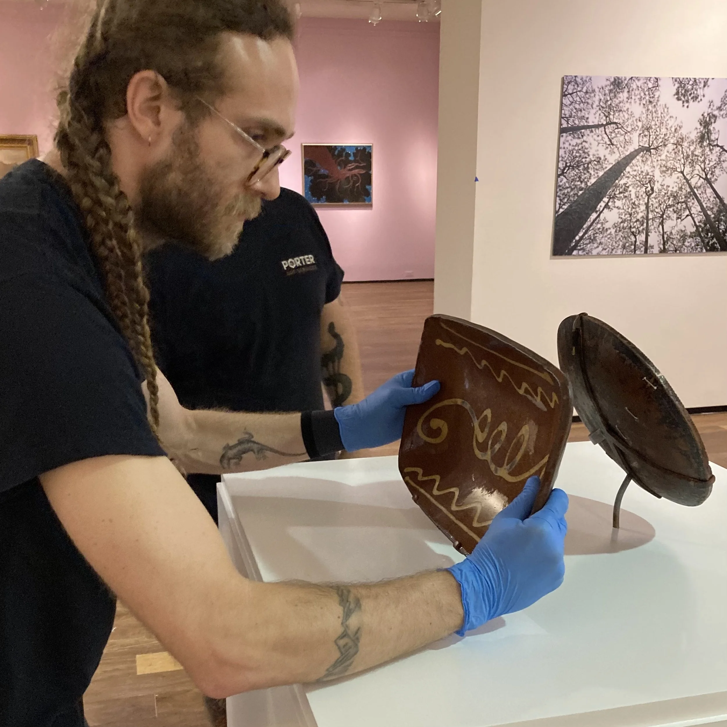 A person in a black t-shirt with tattoos on their arm and beard, wearing blue gloves, is holding a large chocolate cake decorated with yellow and white icing, preparing to serve it at an art gallery or museum with pink and white walls and framed artw
