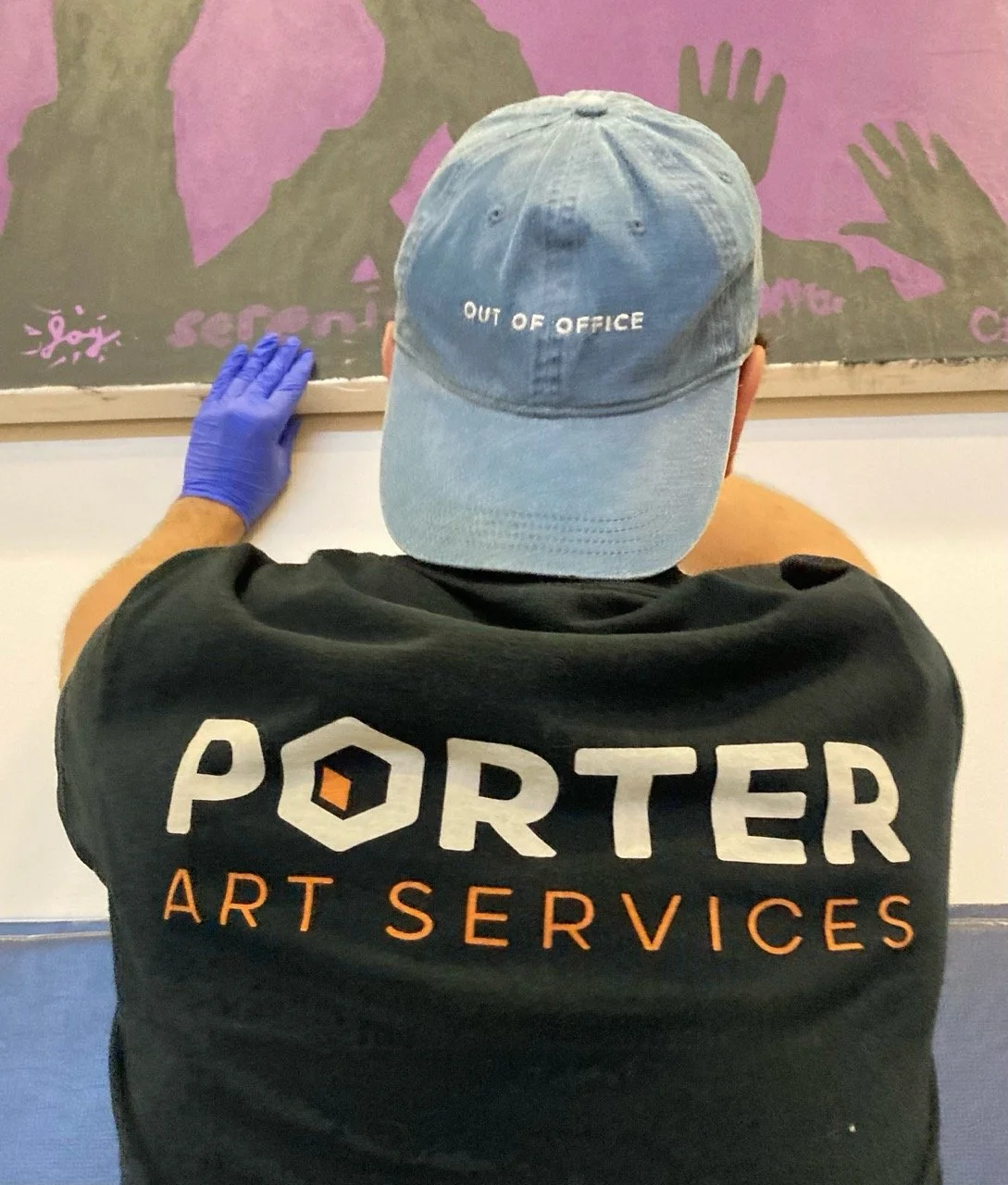 Person wearing a black shirt with orange and white text that reads 'PORTER ART SERVICES' and a light blue cap with 'OUT OF OFFICE' embroidered on it, touching a wall with a purple and brown mural with silhouettes of hands and an eye.