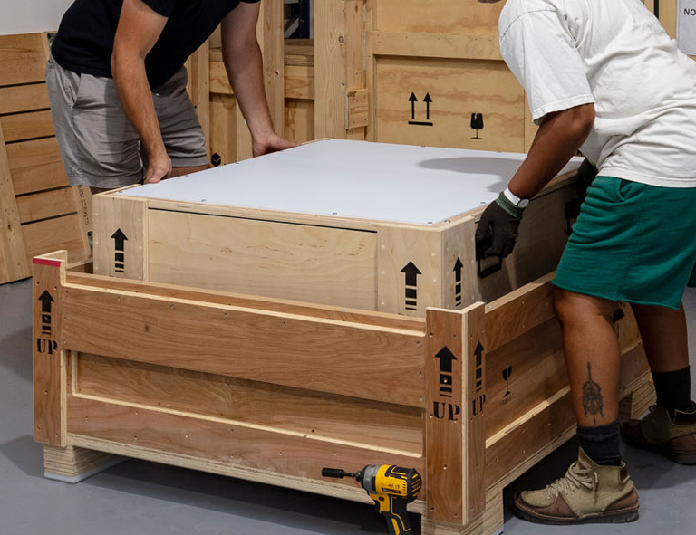 Two people assembling a large wooden crate or enclosed box, with one person wearing gloves and earpieces, in a workshop or storage area.