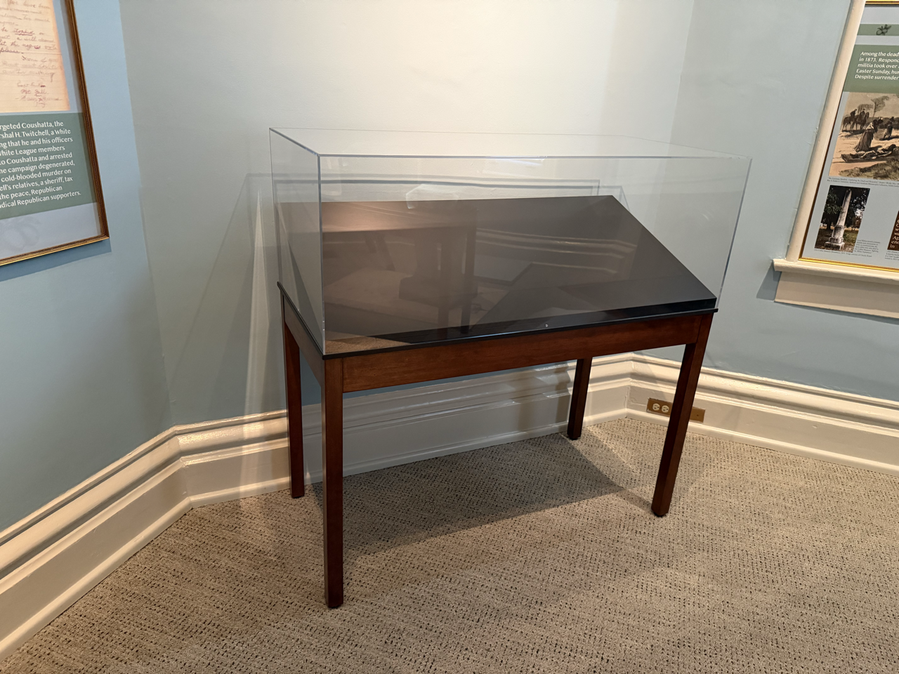 A display case on a wooden table in a room with light blue walls, a framed informational poster on the left, and another framed poster on the right. The display case is empty and has a glass cover.