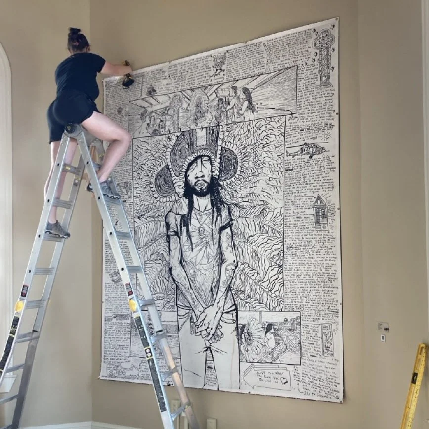 A woman on a ladder drawing a large mural on a wall that features a prominent portrait of a man with dreadlocks and a large hat, surrounded by intricate artwork and text.