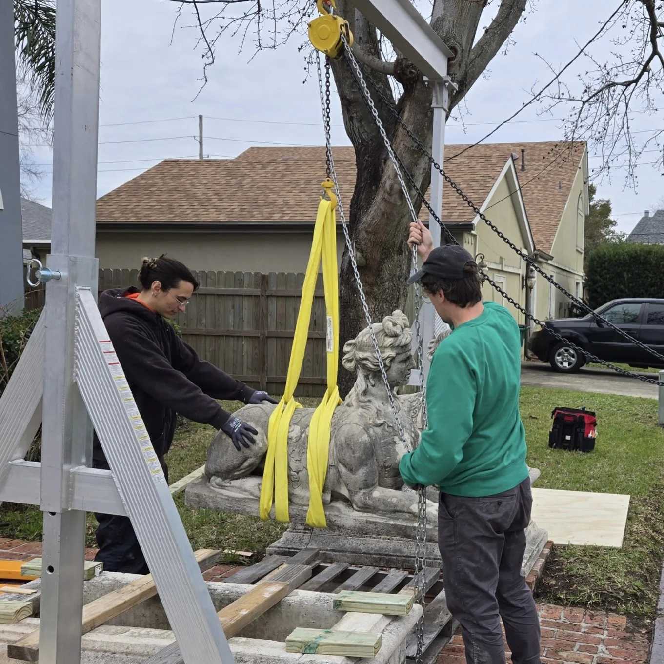 Two workers are carefully lowering a stone lion statue into a grave with yellow straps, chains, and a crane setup in a backyard with aluminum ladder and tools nearby.
