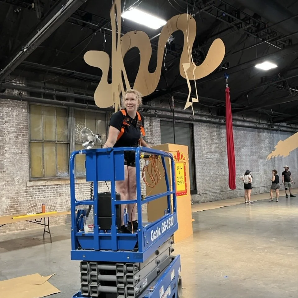 A woman standing on a blue scissor lift inside a building with a brick wall. Hanging above her is a decorative, abstract installation. Three other people are seen in the background near the wall.