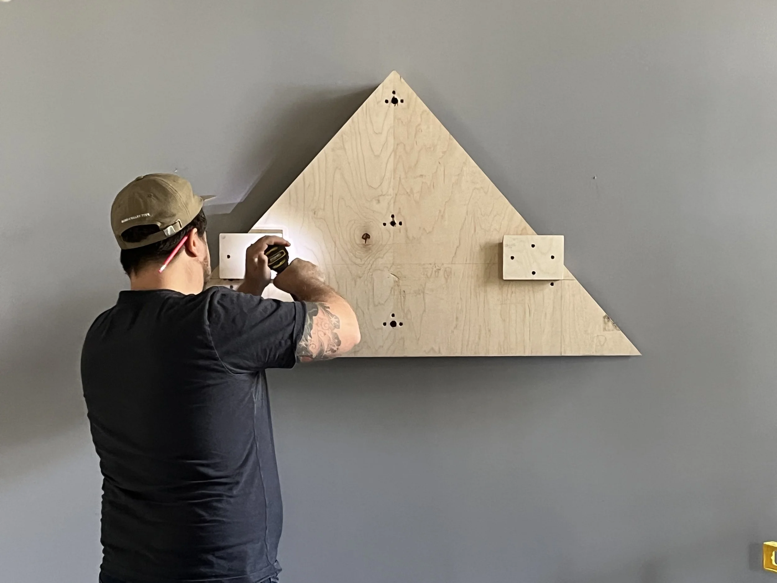 A man wearing a beige cap and black T-shirt using a tape measure on a large wooden triangular structure mounted on a blue wall.