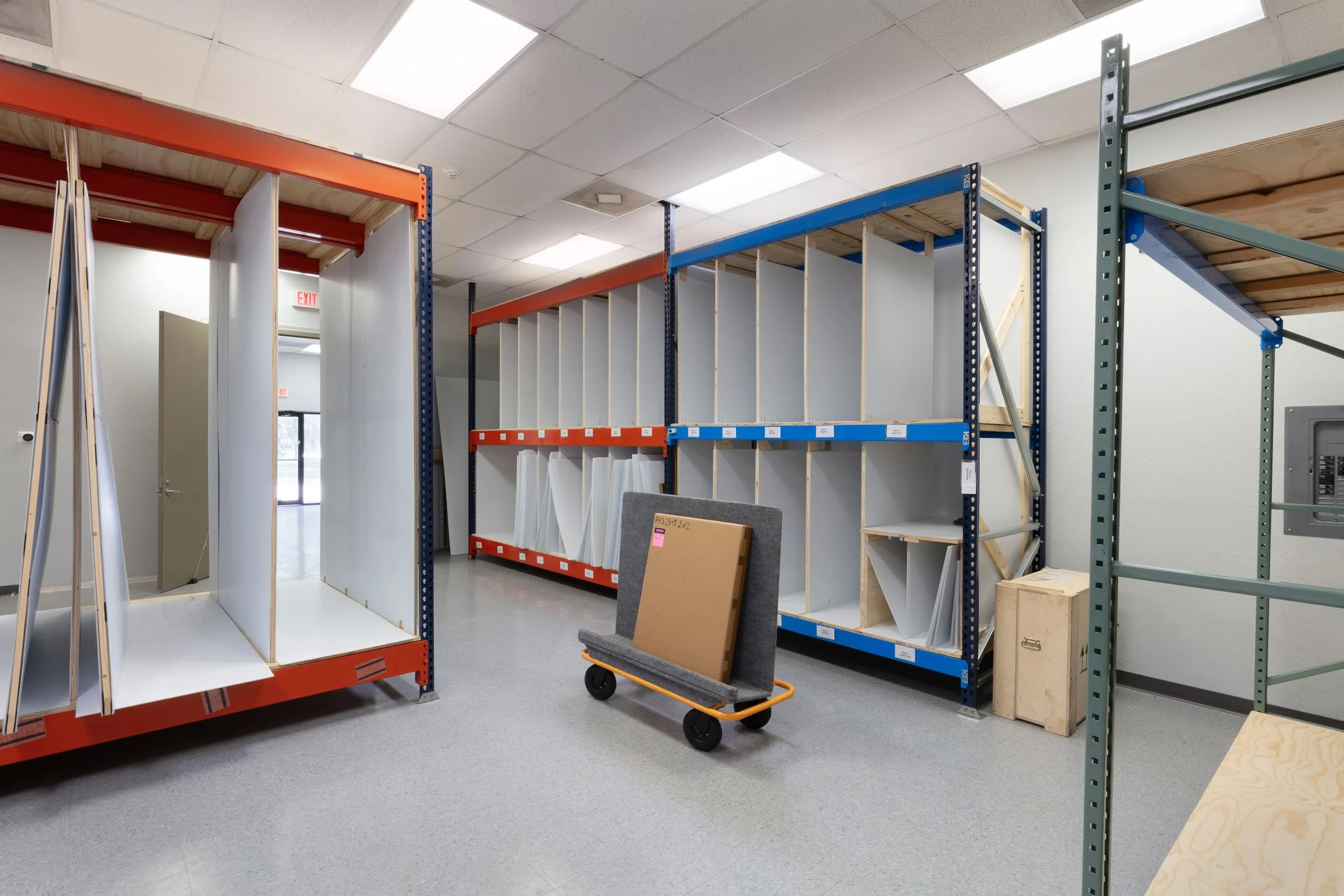 Empty retail store shelving units with plywood and metal frames, a gray cart with a brown box and a gray cushion, and a wooden board on the floor, under a white ceiling with fluorescent lights.