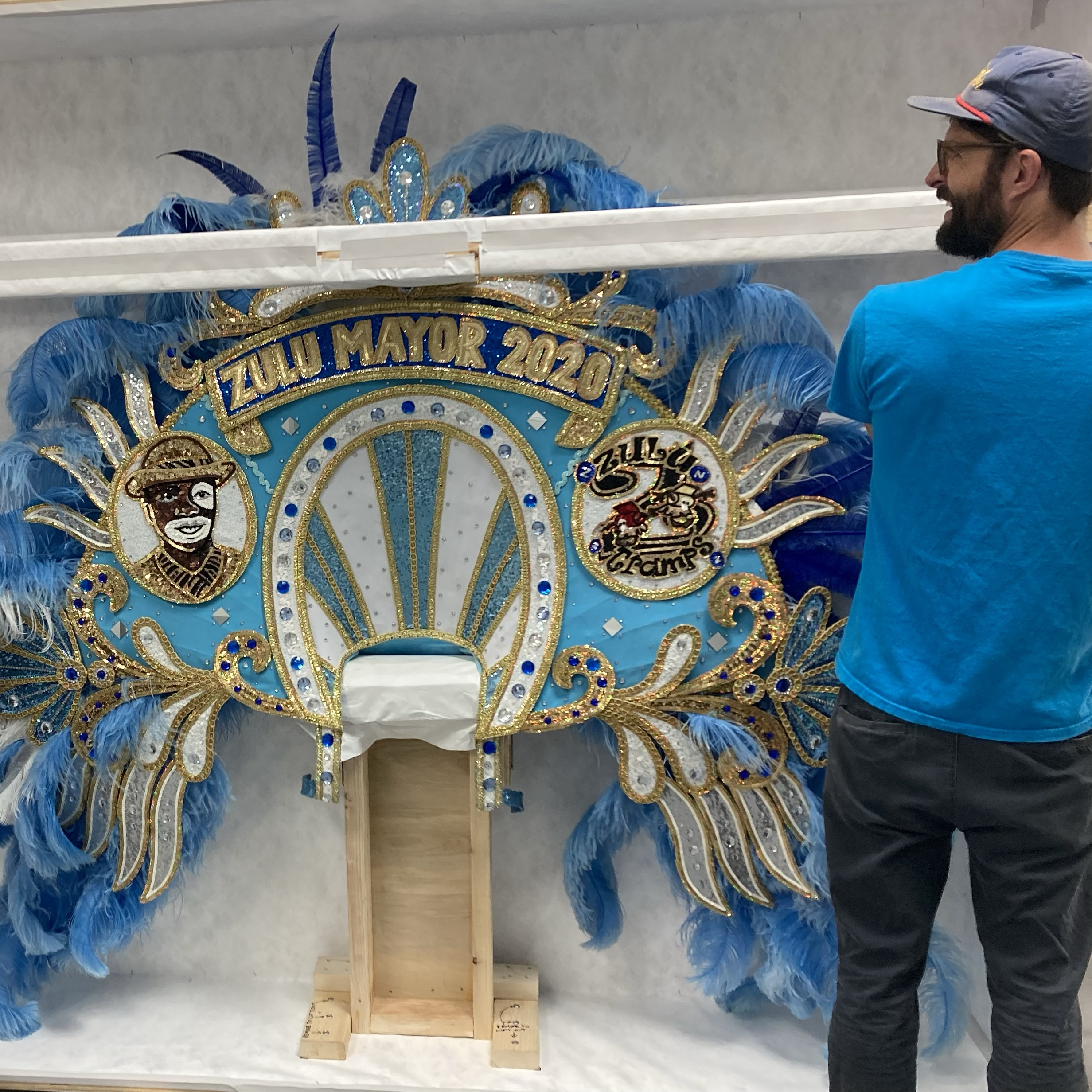 Costume with blue feathers and gold embellishments for a parade or celebration, displayed on a wooden stand, with a man wearing a gray cap in the background.