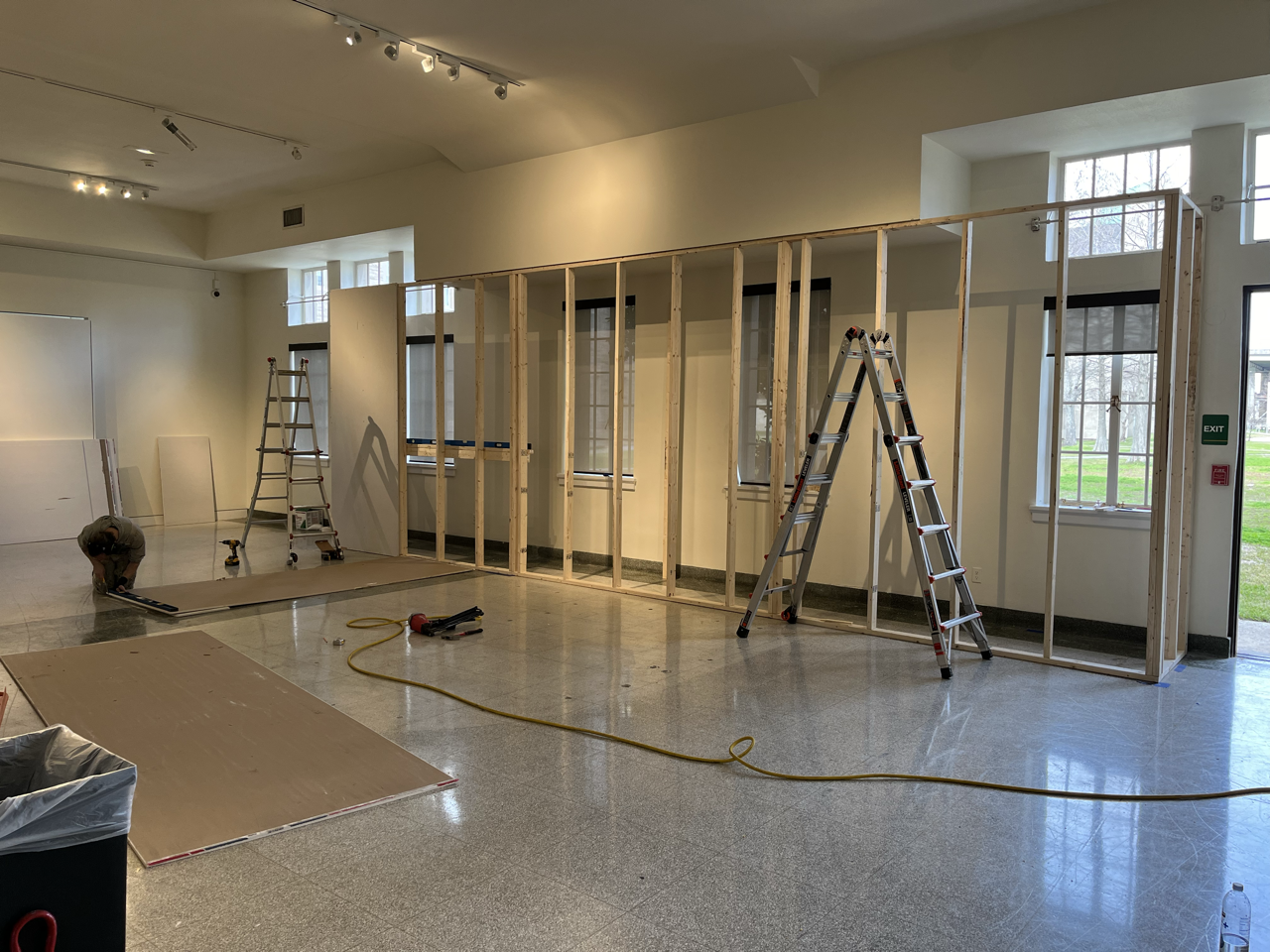 Construction workers are building a wooden frame on an interior wall in a large room with high ceilings and tall windows, using ladders and tools.