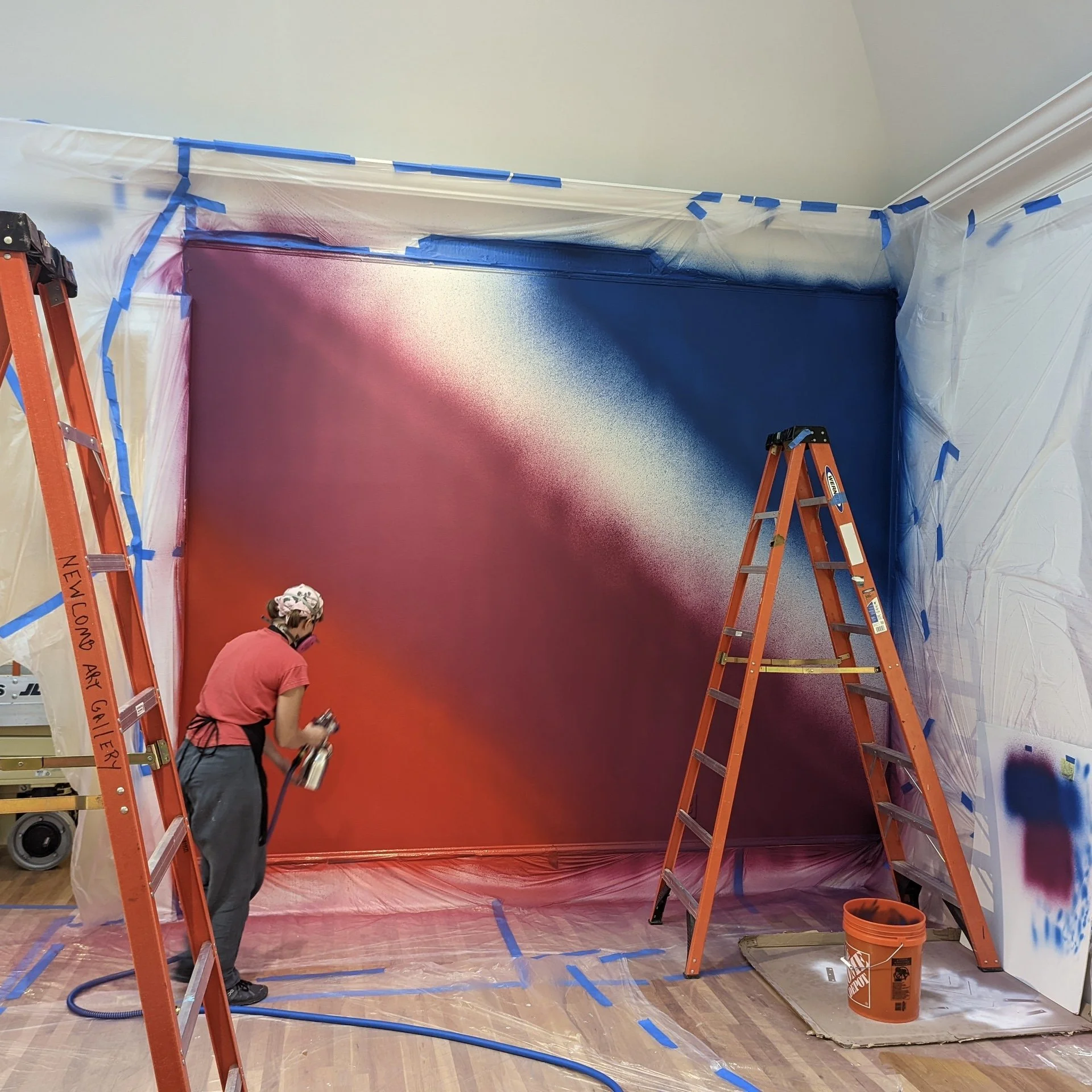 Person spray painting a wall with a gradient red to silver effect, surrounded by plastic sheeting and ladders.