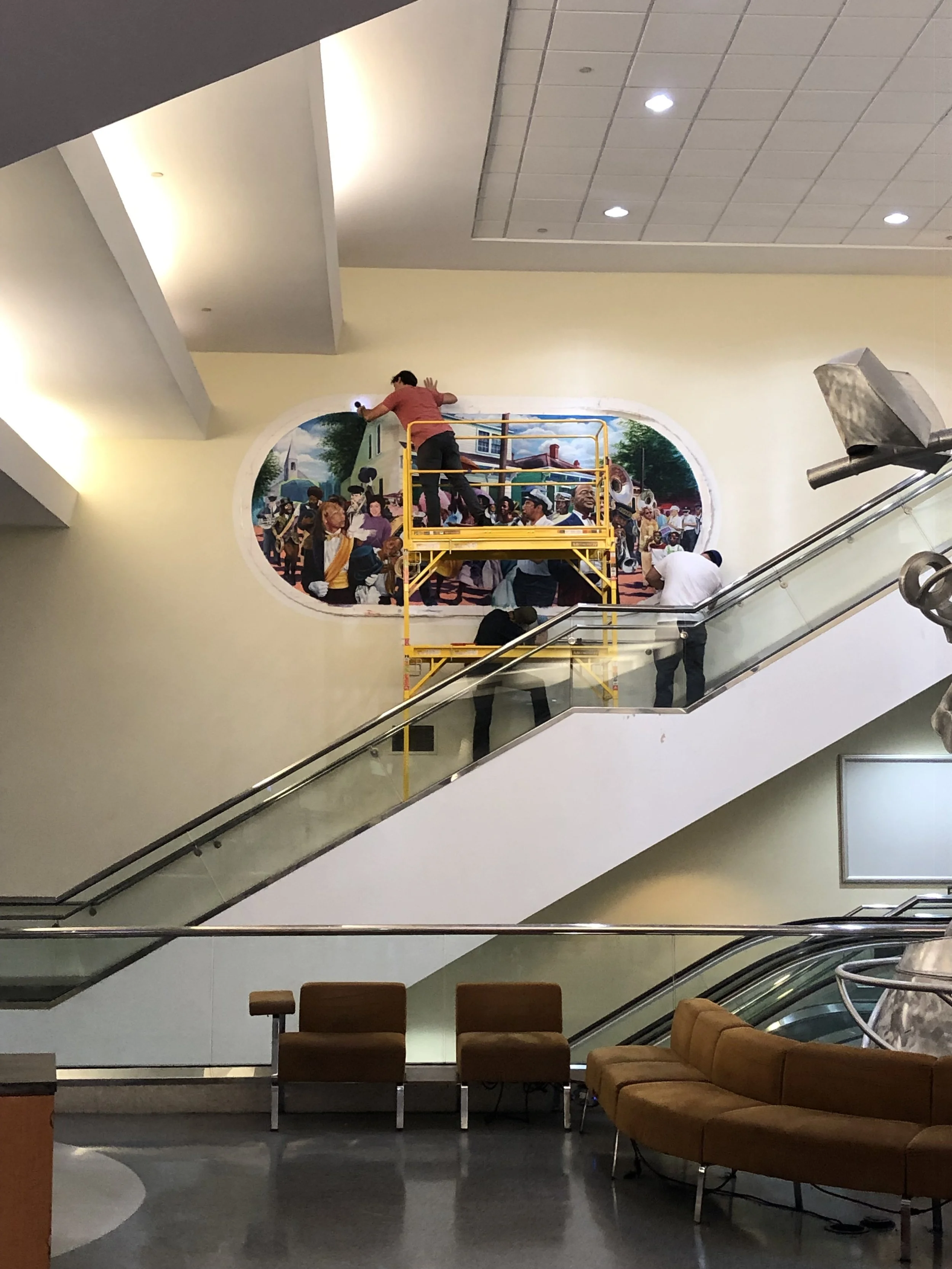Interior of a shopping mall or airport with a mural of a festival scene on the wall. Two people are on a yellow scaffold working on the mural. There are escalators, chairs, and lobby furniture visible.