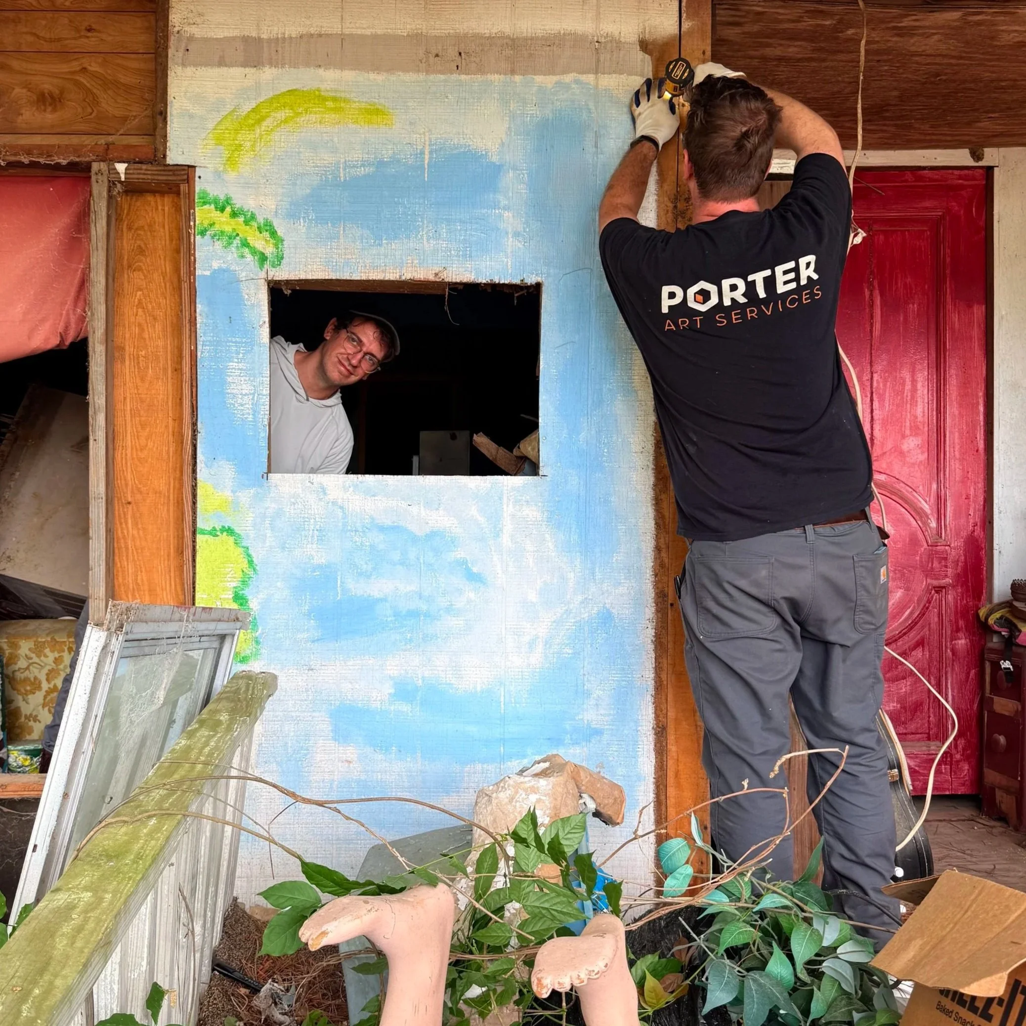 Two men working on an art project on a wall with partially painted and unpainted areas, one wearing glasses and a hat peeking through a window, the other using a drill, in a room with wood paneling and plants in the foreground.