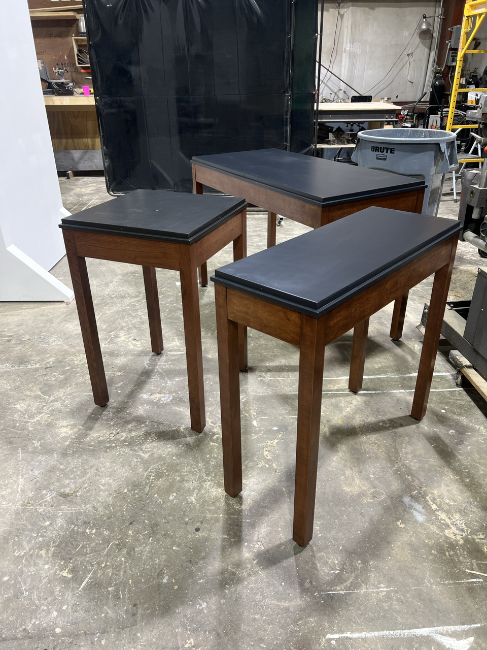 Three wooden tables with black tops arranged in a workshop or storage area, with various tools and equipment in the background.