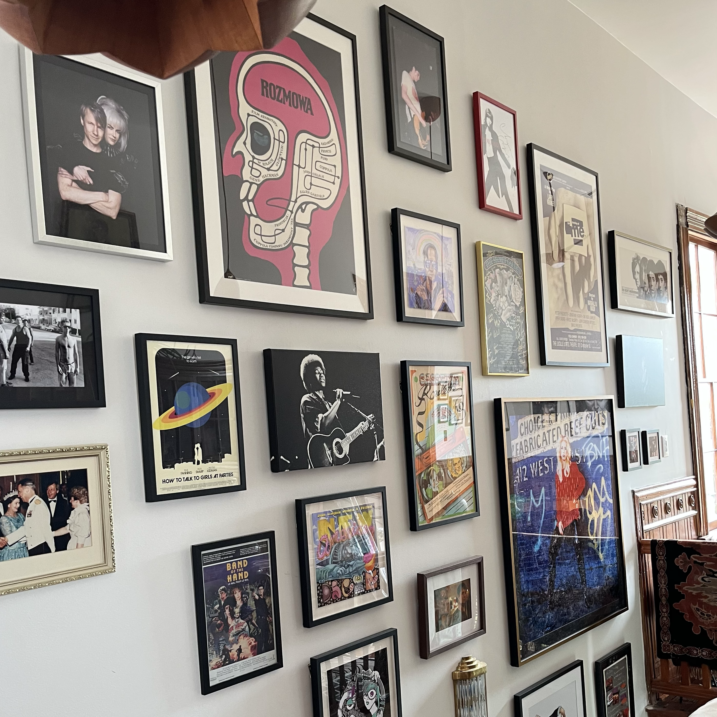 A wall with various framed posters and artwork, including a colorful poster of a skull with text, a black-and-white photo of two people, and other artistic prints in different sizes and frame styles.