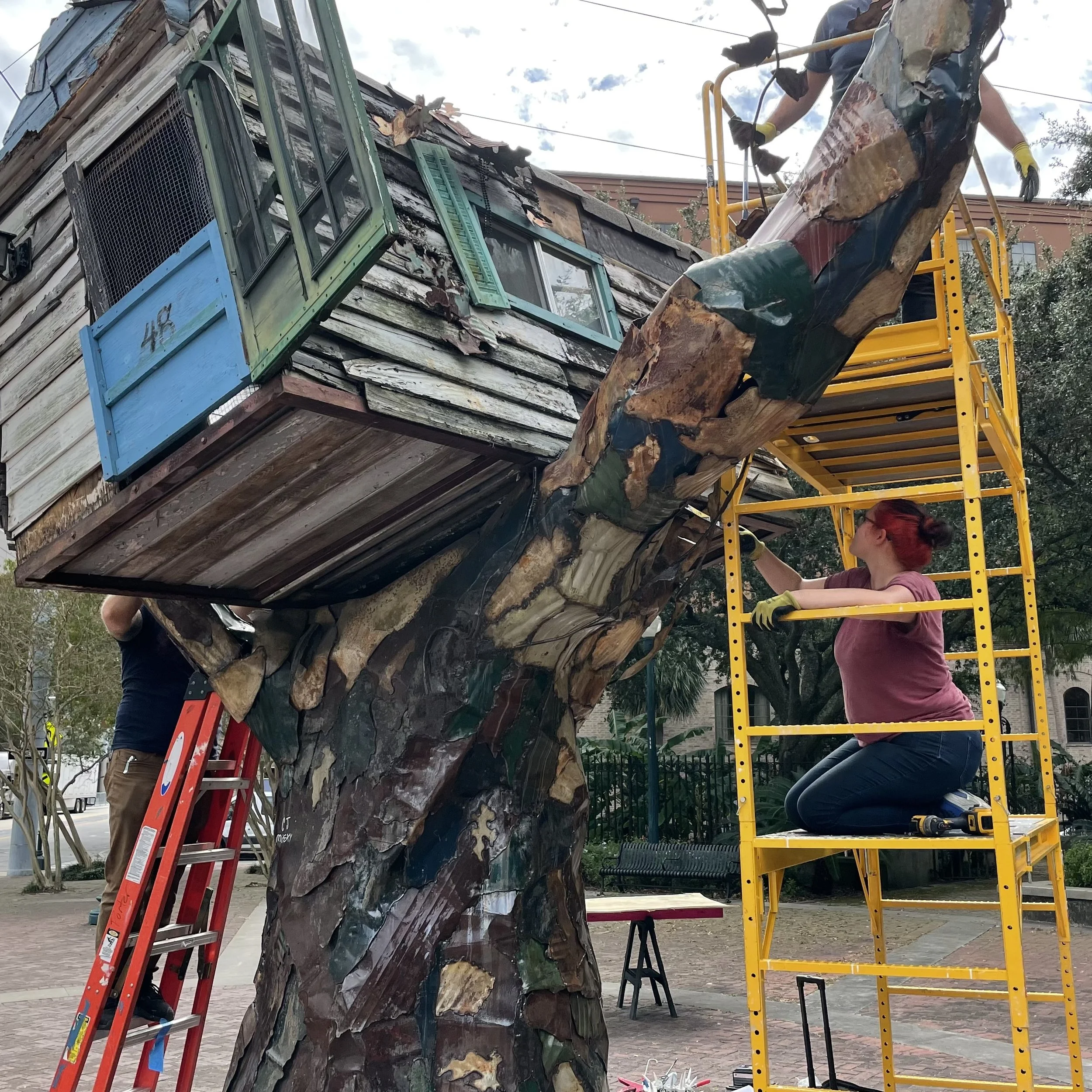 A person working on a large animal sculpture in an outdoor setting with scaffolding, ladders, and tools.