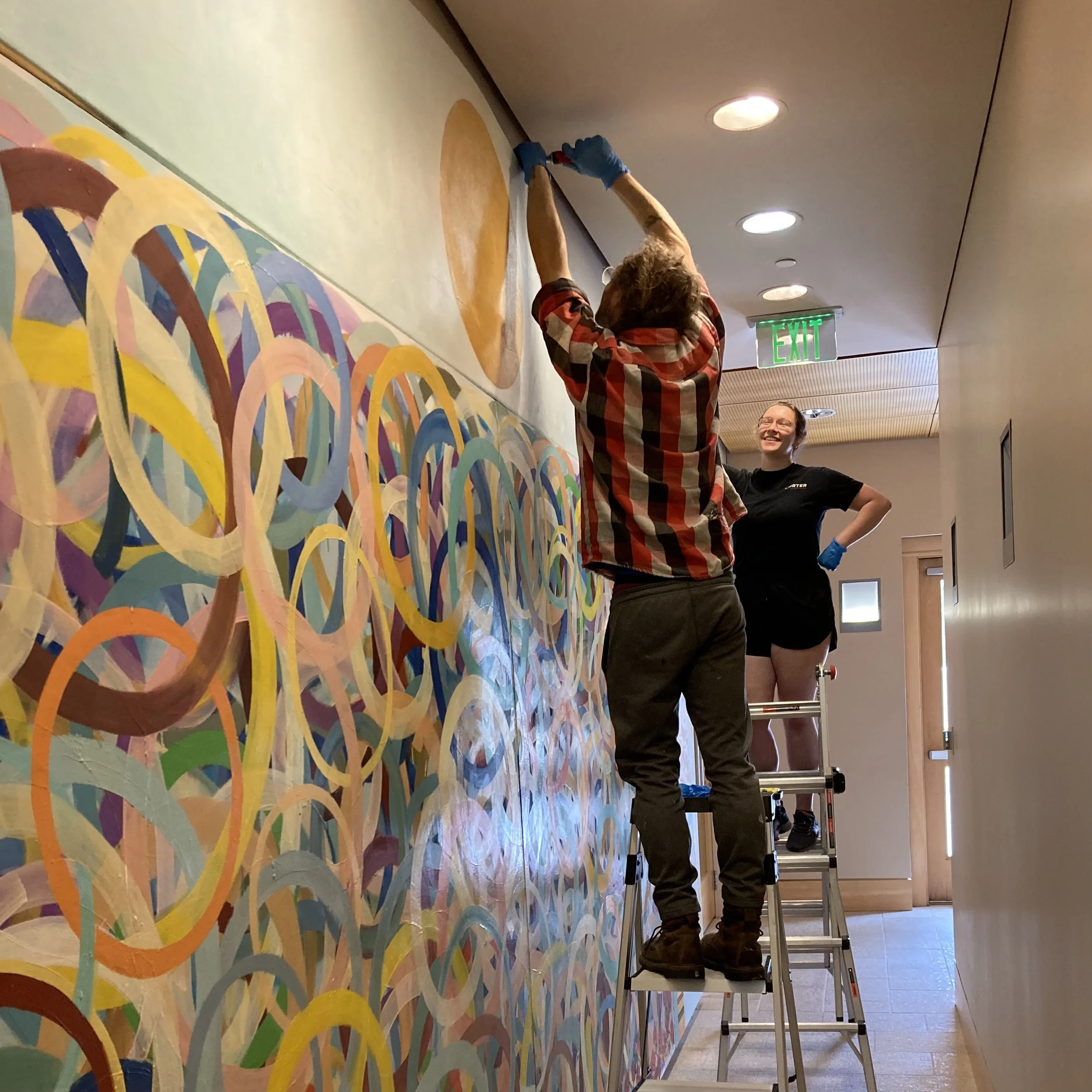 People installing or painting a large, colorful, abstract mural on a wall in an indoor space, with a woman standing nearby smiling and another person on a ladder working on the mural.
