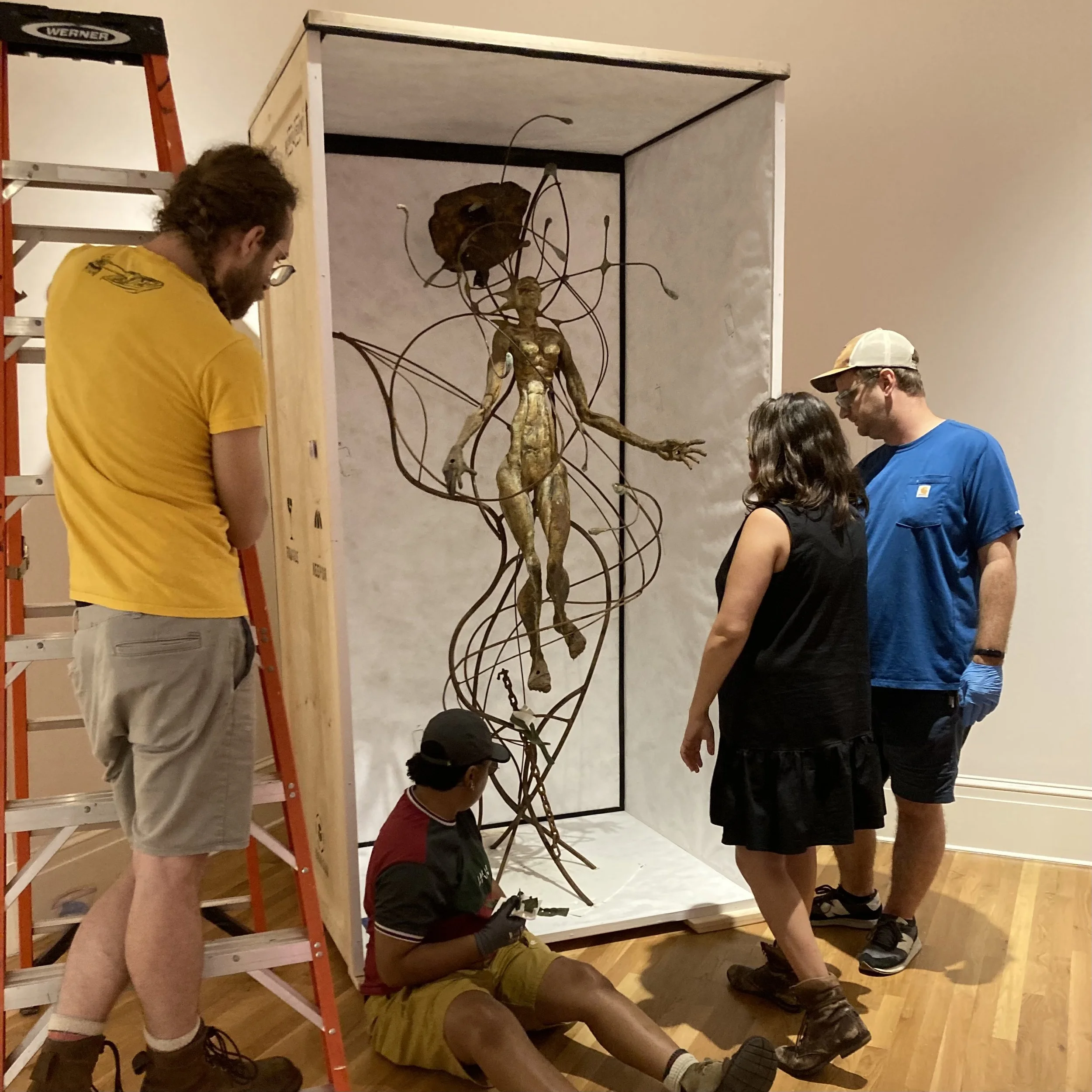 Group of people examining and working on a sculpture of a human figure with interconnected metal wires, inside an art gallery or museum setting.