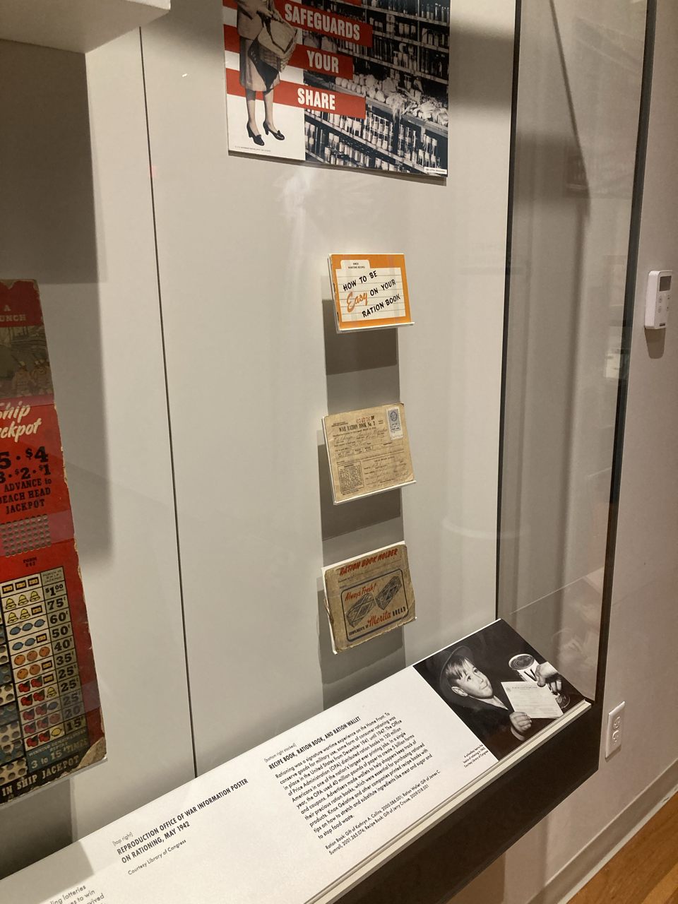 Display case with historical booklets and an informational poster about wartime rationing, featuring a black-and-white photo of a young boy eating with a spoon.