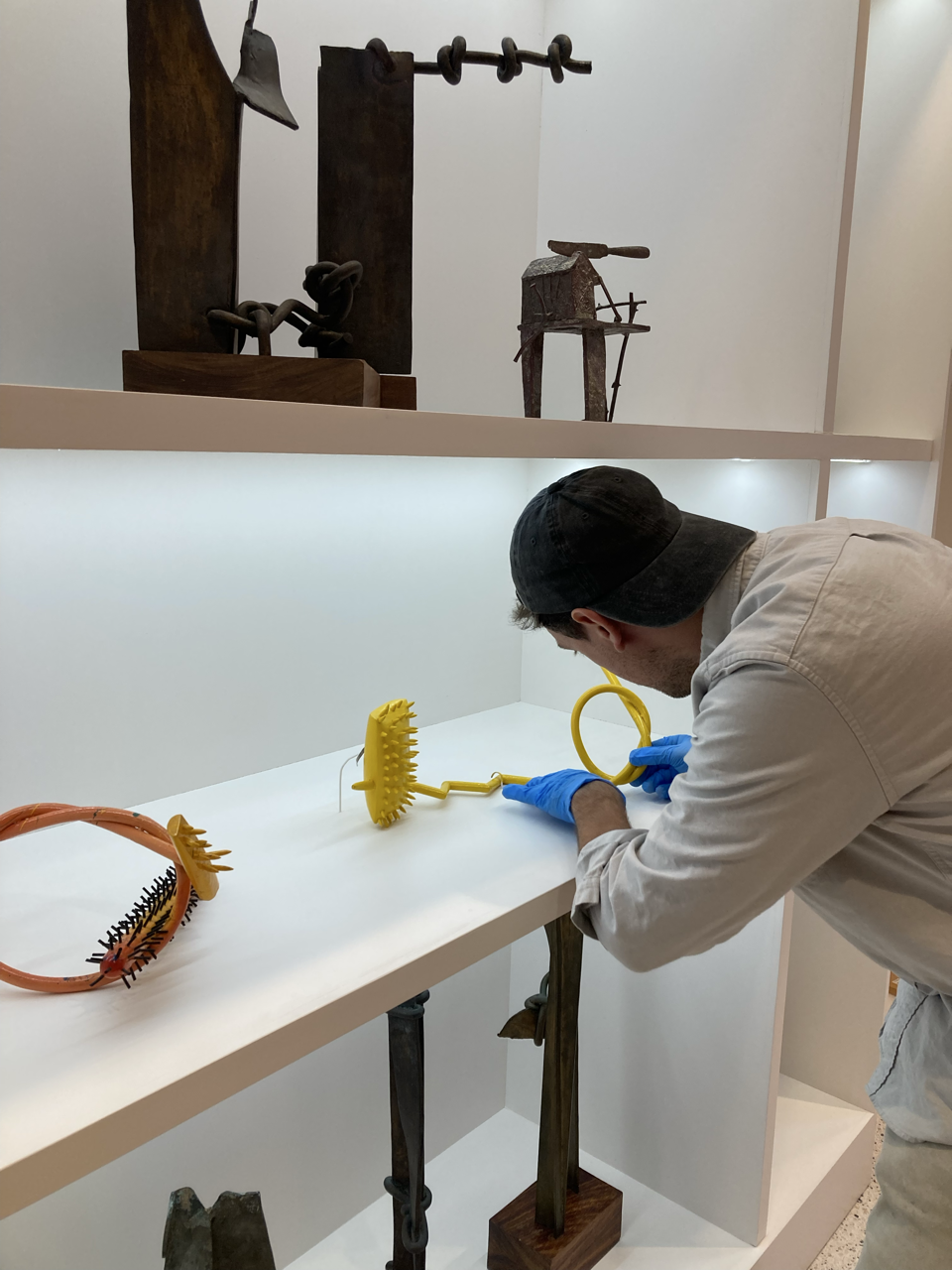 A person in a white shirt, black cap, and blue gloves is adjusting or examining modern artistic sculptures on a white display shelf. The sculptures are colorful and unique, resembling unconventional, artistic forms.