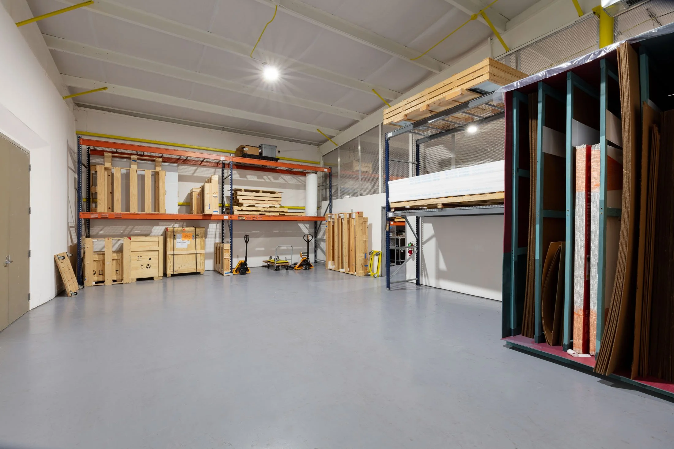 Inside a warehouse or storage unit with metal shelving filled with wooden pallets, boxes, and construction materials. The concrete floor is clear, and the ceiling has exposed beams with a single light.