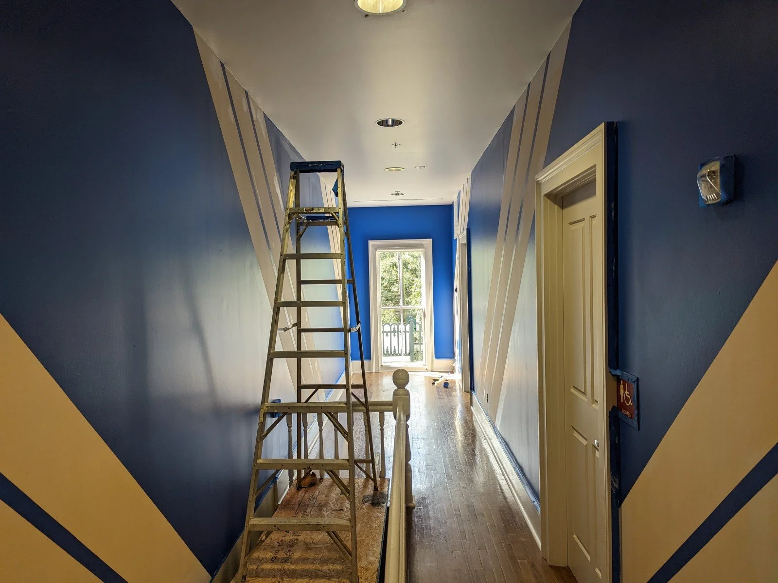 Hallway with blue and beige walls, wood flooring, a ladder, and a window at the end of the hall