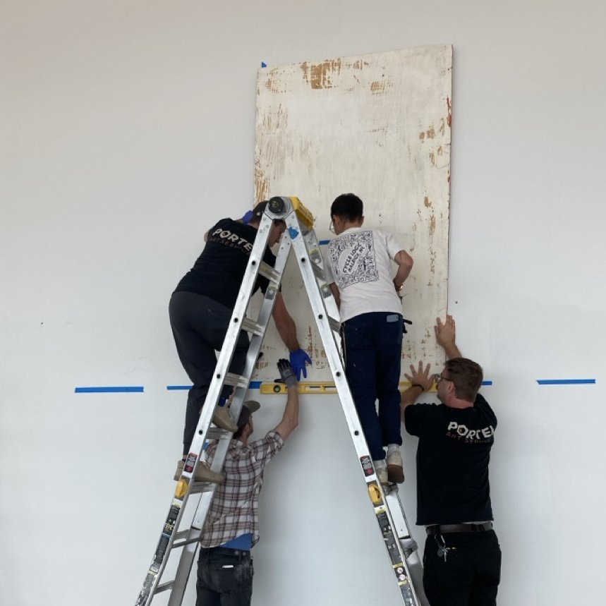 Four people working together to hang a large canvas or painting on a white wall. One person is on a ladder, while two others stabilize the artwork from below. The fourth person is holding the ladder for support.