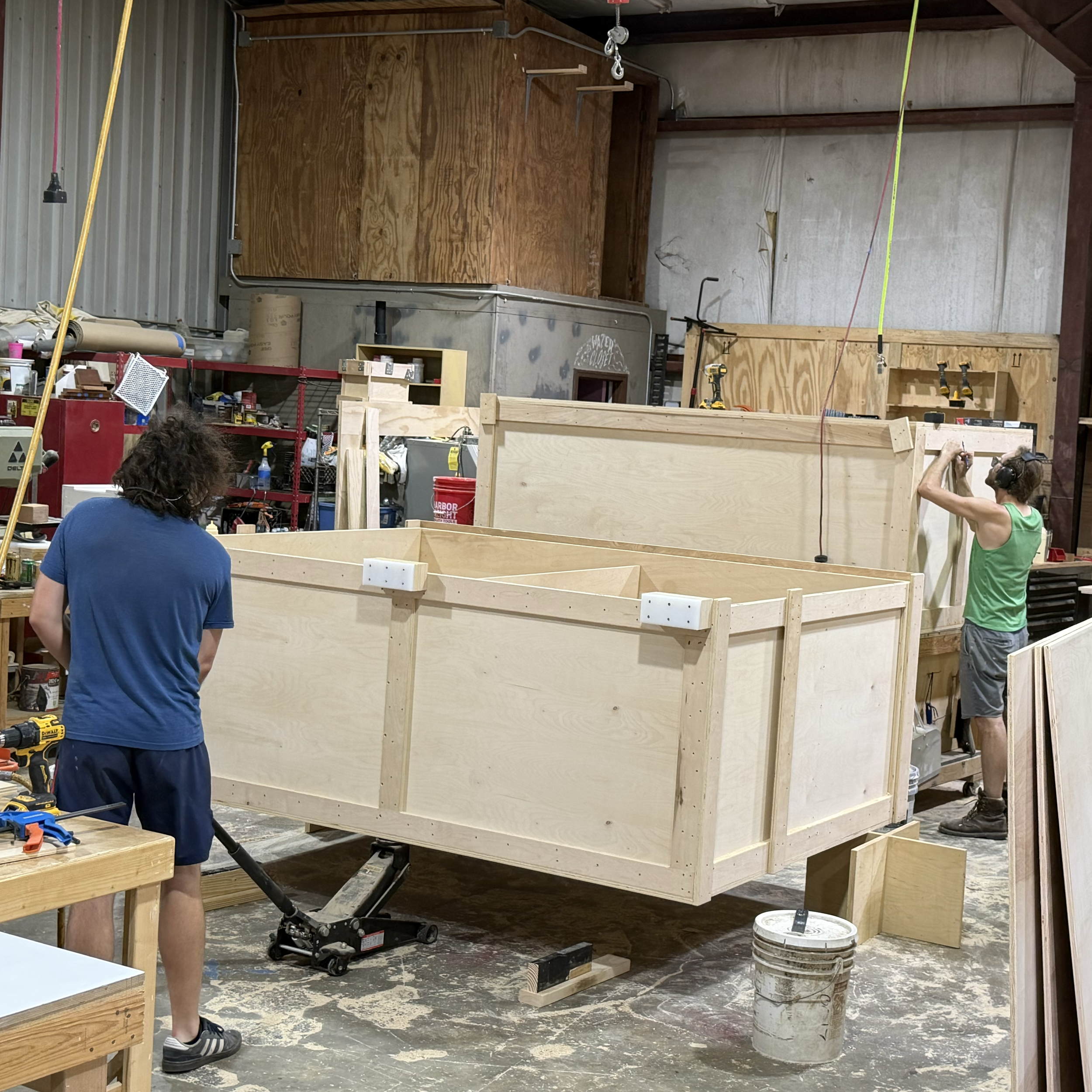 Construction workshop with wooden frame and tools, two workers installing a wooden structure, shelves, electrical wires, and a wheelchair lift in a workshop.