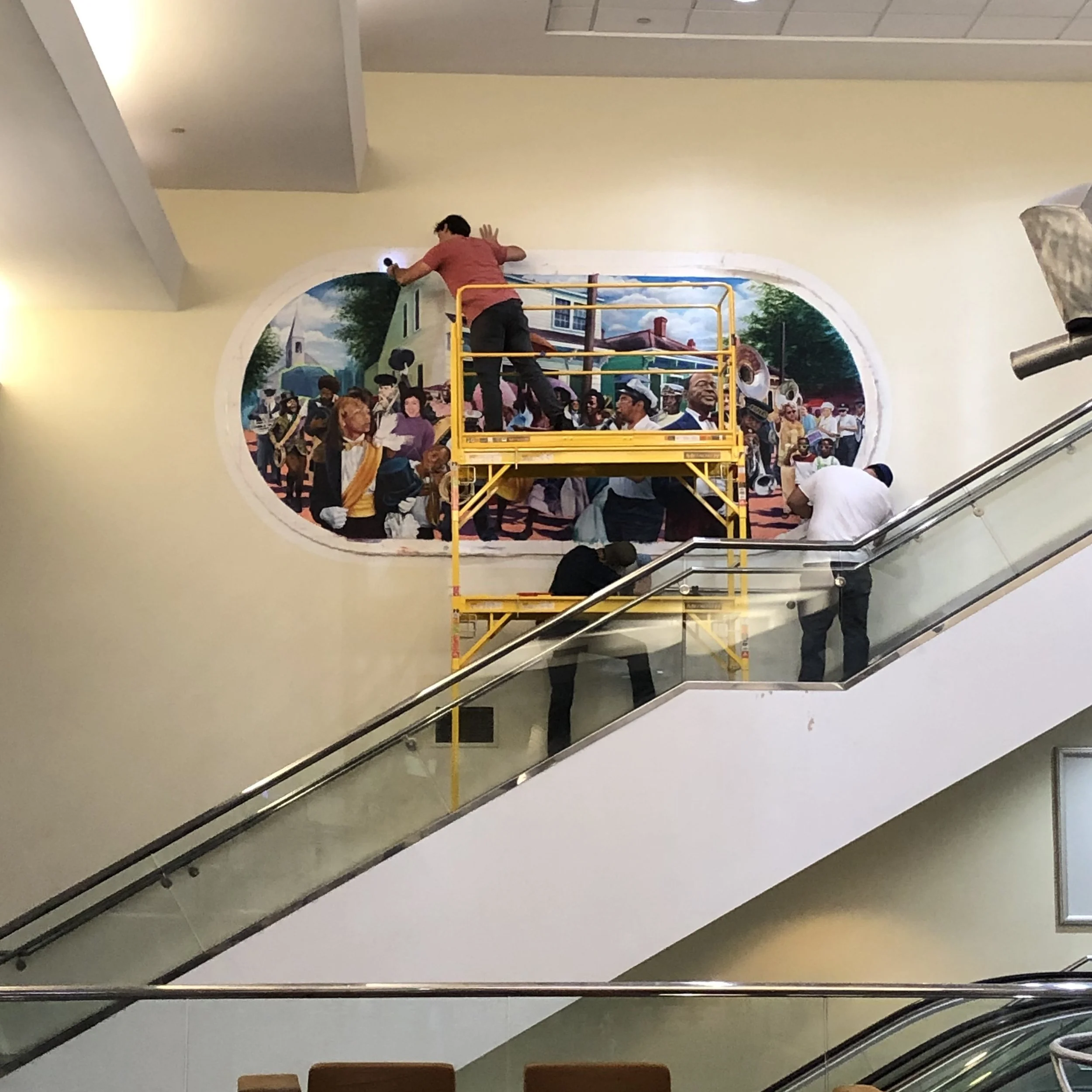 A person in a red shirt is standing on a yellow maintenance lift, painting or working on a mural window on a wall. The mural depicts a countryside scene with trees, a house, and a church steeple, viewed through a circular opening in the wall. There a