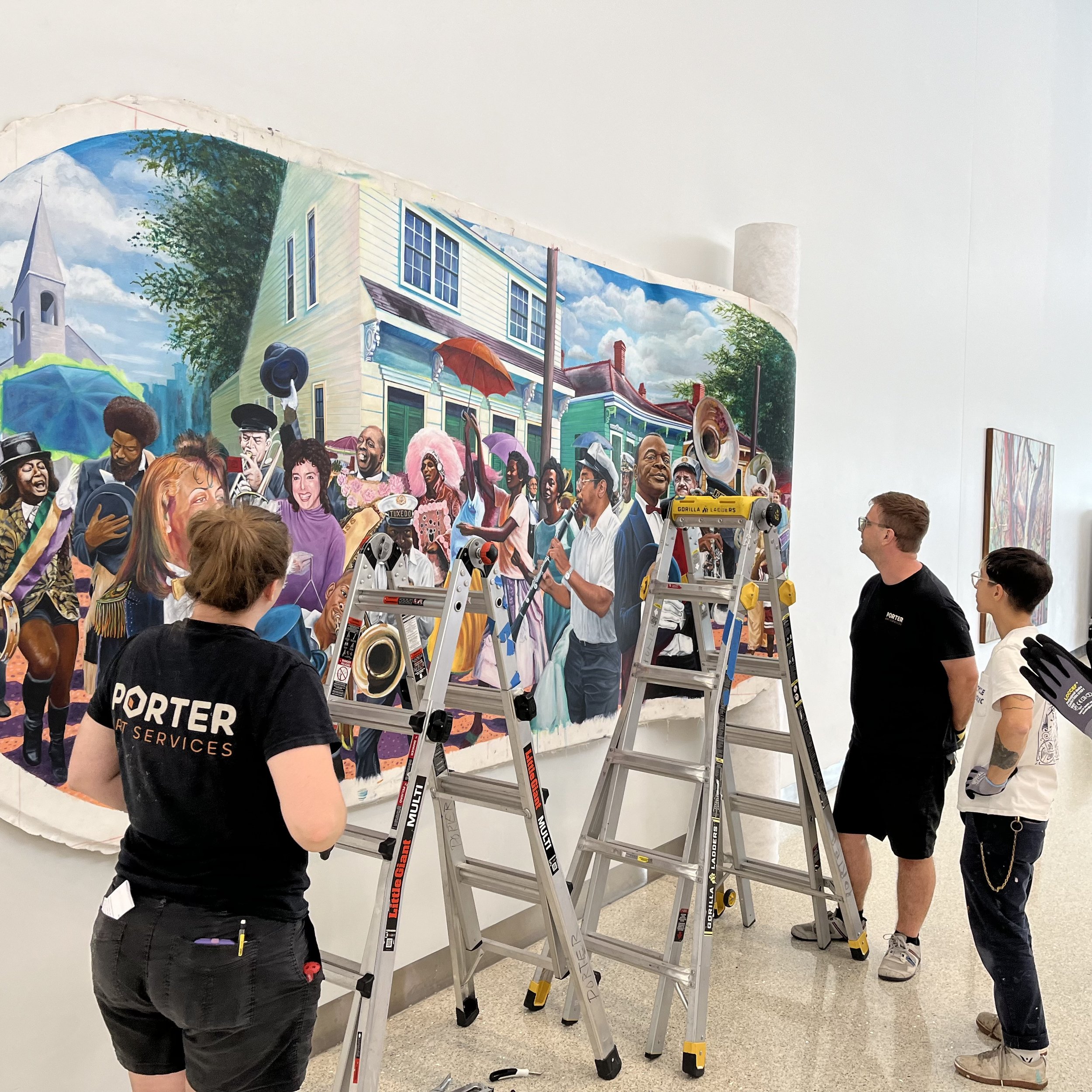A mural depicts a lively street scene with a crowd of people, some holding umbrellas, inside a gallery or museum. There are ladders and tools set up, indicating the mural is being painted or worked on.