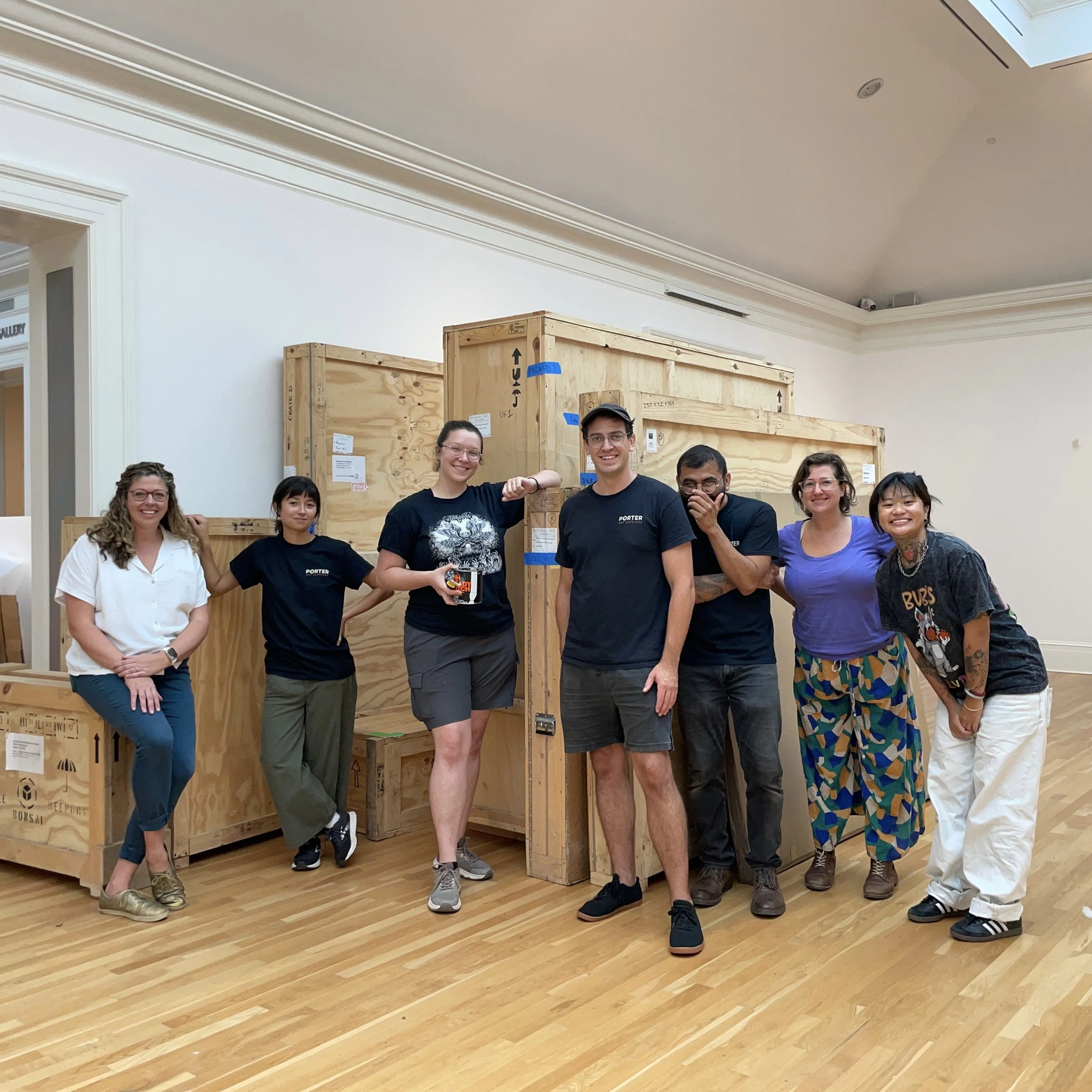 A group of seven people standing indoors in front of large wooden crates, smiling and posing for a photo. The room has white walls and wooden flooring.