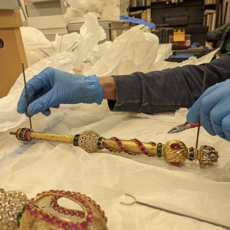 A person wearing a gray long sleeve shirt and blue gloves working on an ornate gold and jewel-encrusted scepter or decorative staff, with multiple tools and paper in a workshop or conservation studio.