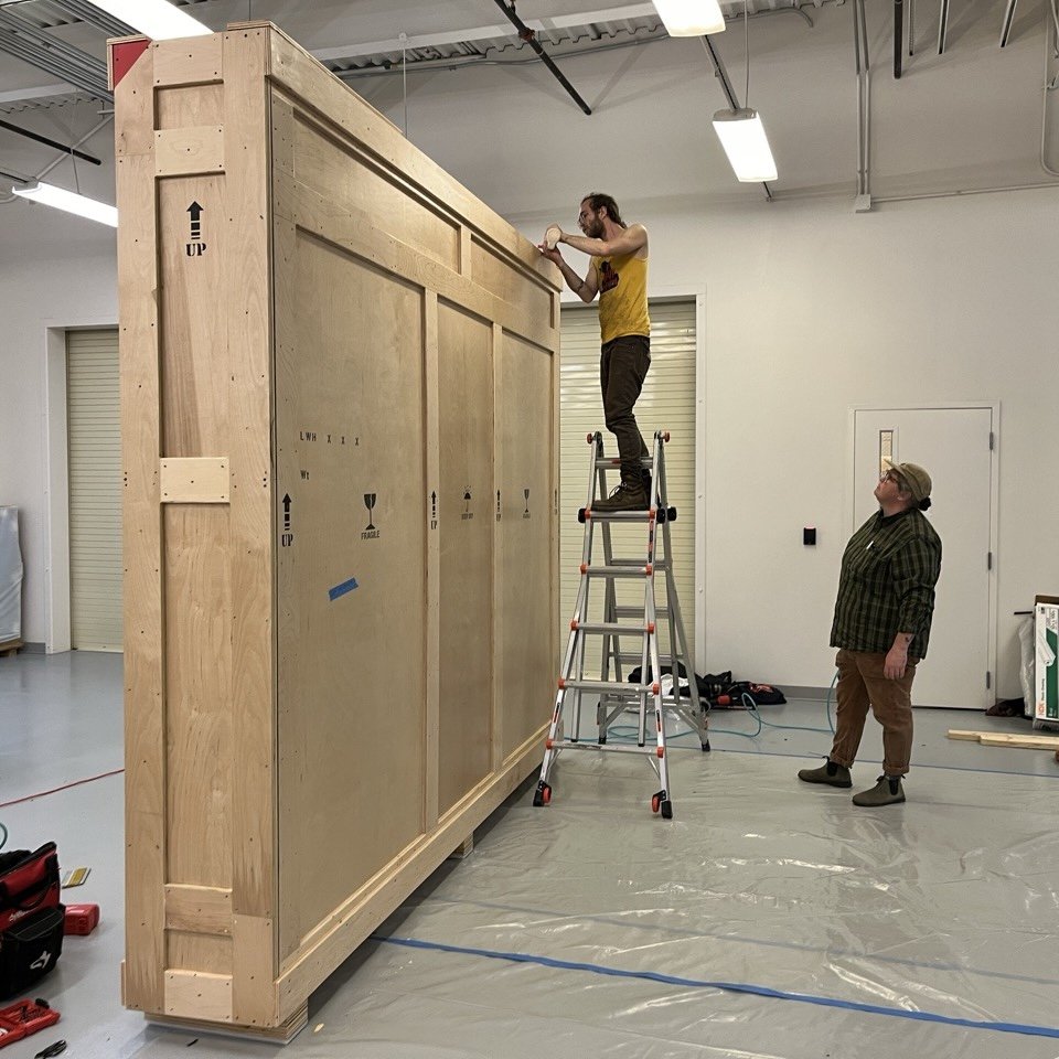 Two people working on assembling a large wooden crate inside a room with a ladder. One person is standing on the ladder using a tool, and the other person is standing on the floor watching.