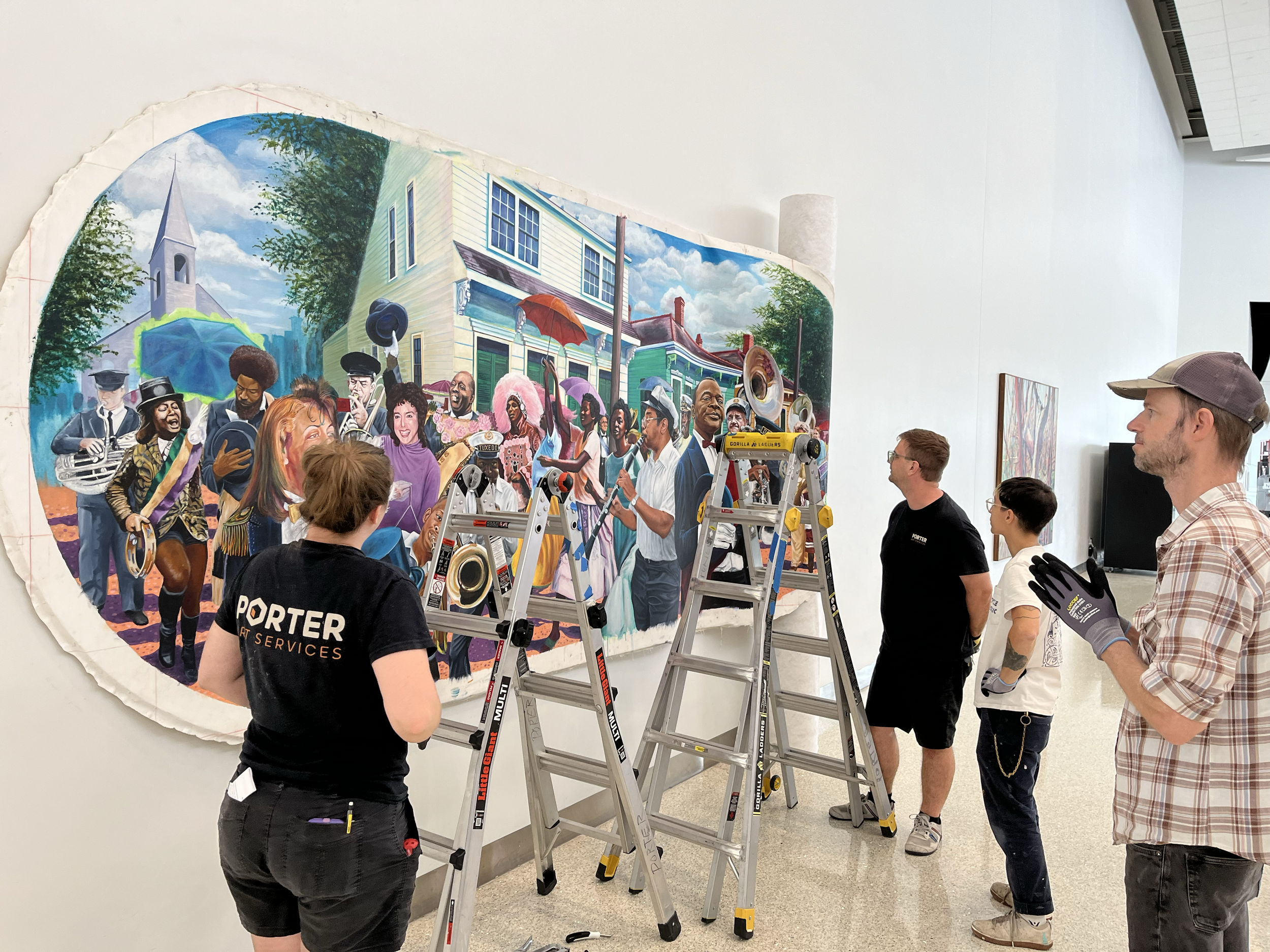 People standing in front of a large colorful mural inside an art gallery, with some using ladders to work on it.