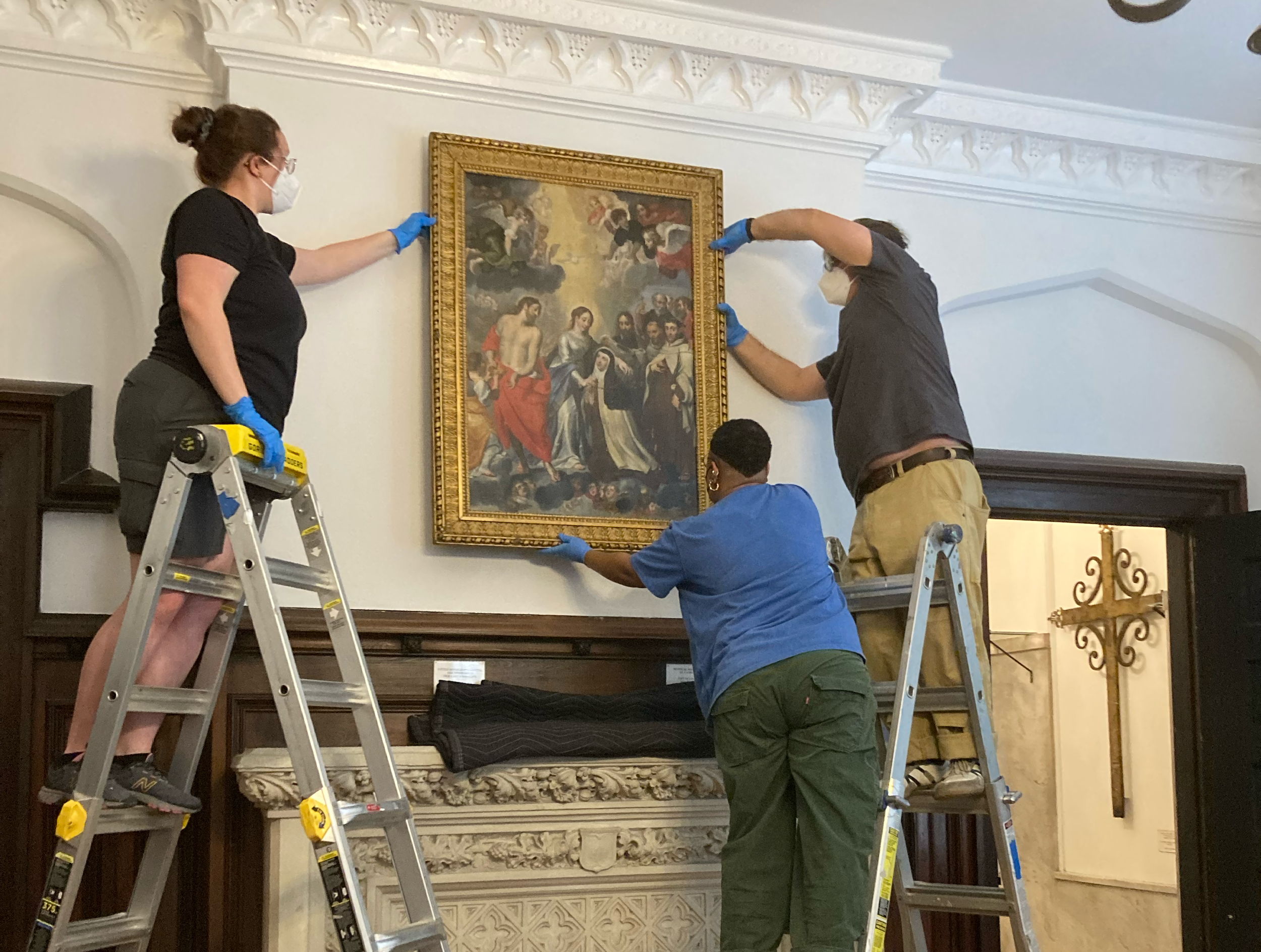 Four museum workers are hanging a large framed painting on the wall, with two standing on step ladders and one on the ground helping. They are wearing masks and blue gloves, in an ornate room with decorative crown molding and a detailed fireplace man
