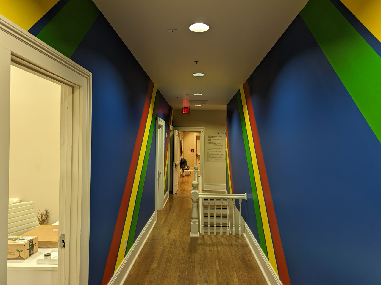 Colorful hallway with rainbow diagonal stripes on navy blue walls, wood flooring, white trim, and a stairway with white railing at the end.