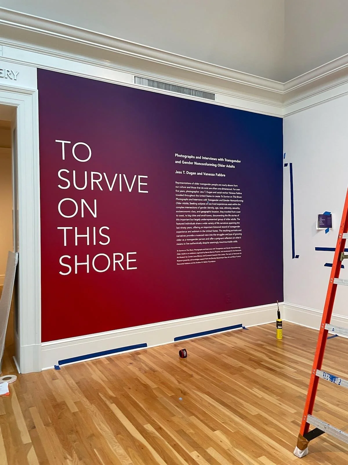 A large exhibition wall with a gradient background from purple to red, displaying text that reads, 'TO SURVIVE ON THIS SHORE', along with additional detailed information about an art project or exhibition, and a ladder and tools are present in the ro