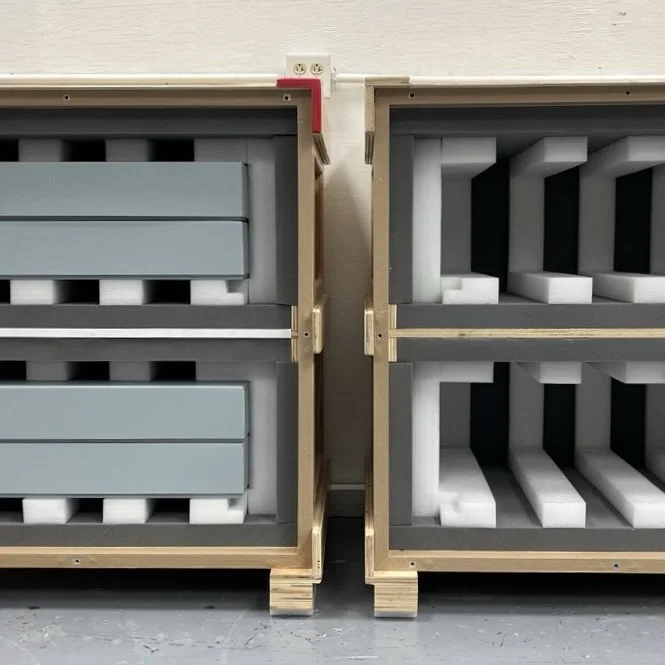 Two wooden crates with foam inserts for packaging or storing 2D artworks, placed side by side against a plain wall.