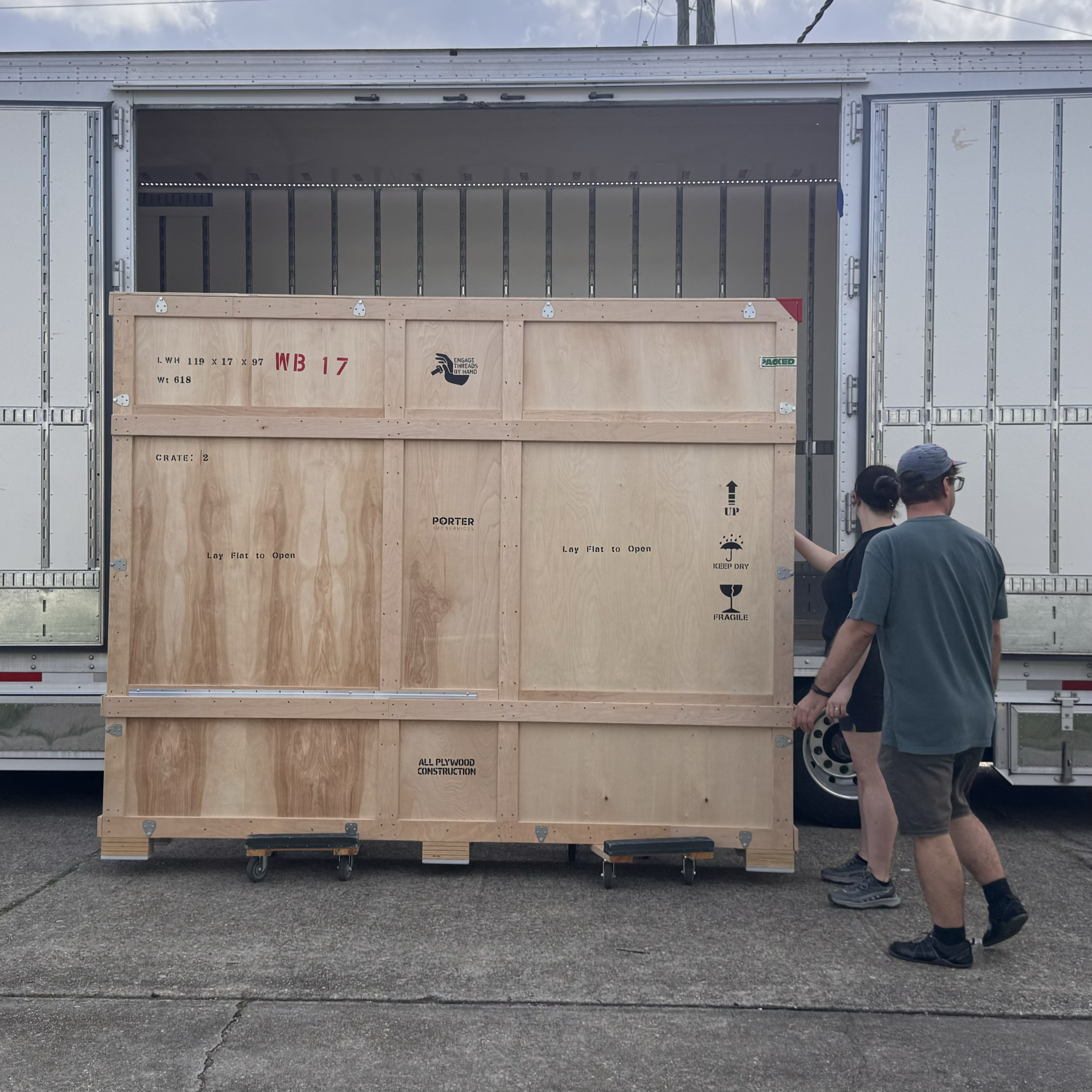 A large wooden crate and a large wooden box on wheels in front of a truck trailer, with people standing nearby. The crate has markings including 'WB 17,' 'Engage Threads by Hand,' and 'Porter.' The box has markings including 'Lay Flat to Open,' 'Keep