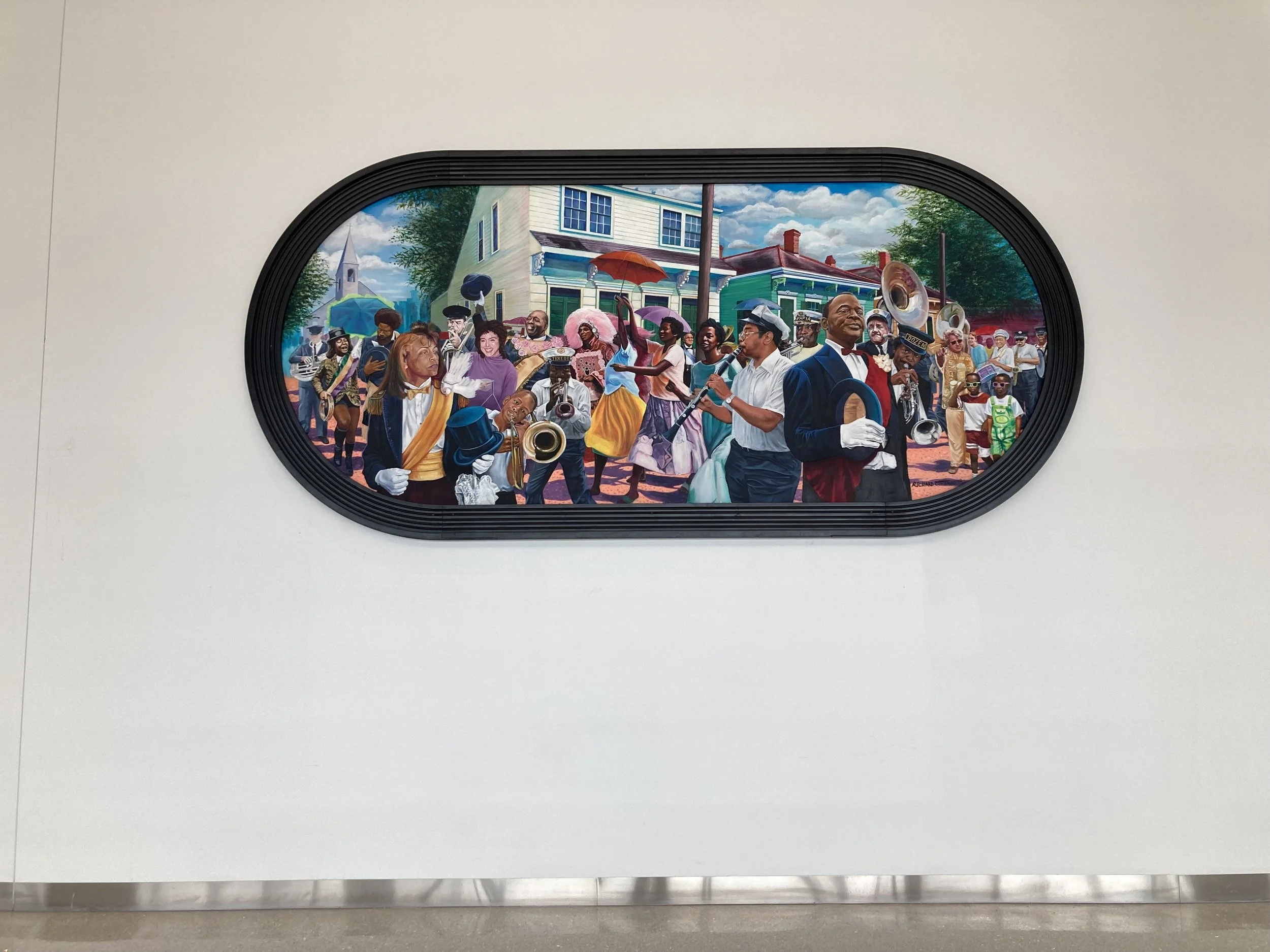 Colorful mural of a street scene with people dancing and playing musical instruments, with houses, trees, and a church in the background. The mural is framed in an oval shape.
