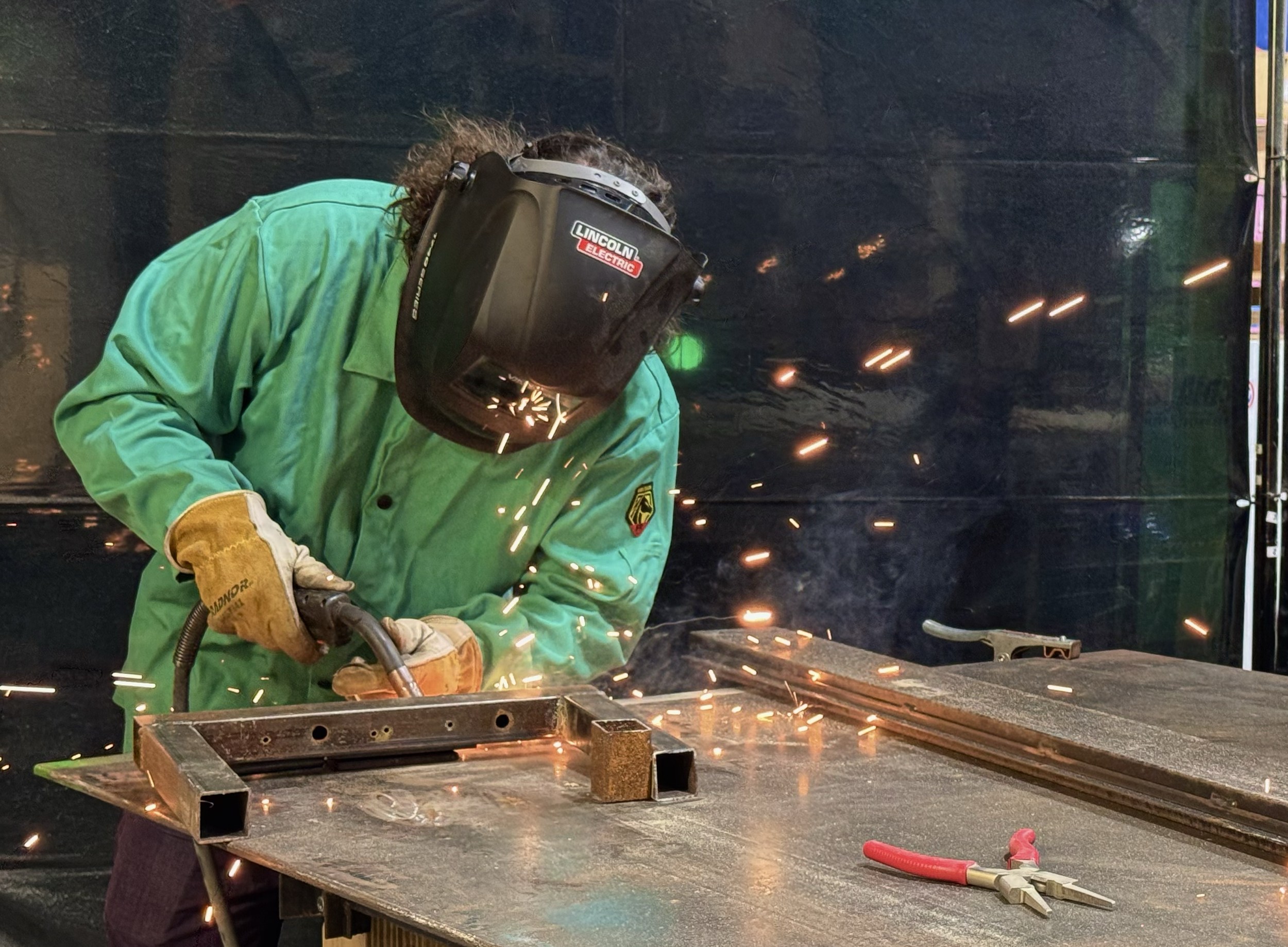 A person welding metal at a workshop, wearing a green protective jacket, welding helmet, and gloves, with sparks flying around.