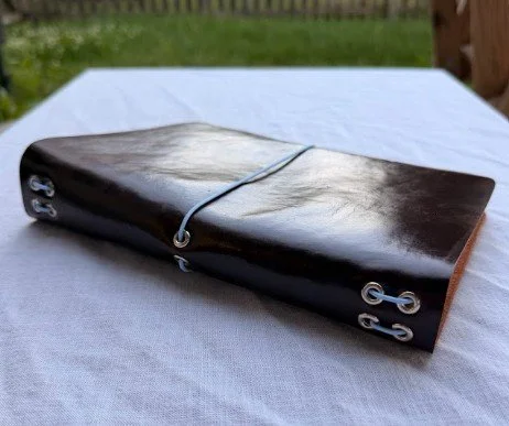 A closed, dark-colored leather-bound journal with metal rings and a paperclip fastened to the front cover, placed on a white surface outdoors.