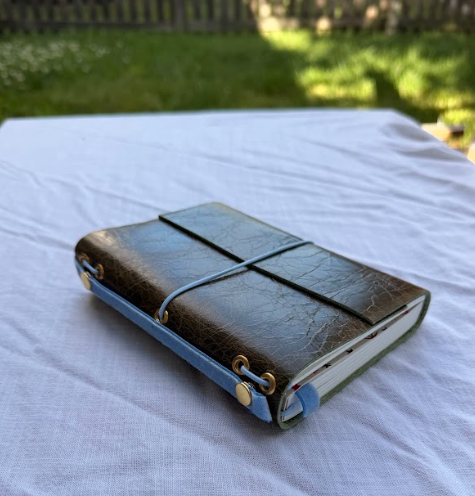 A closed green leather-bound journal with a rubber band around it, resting on a white cloth outdoors.